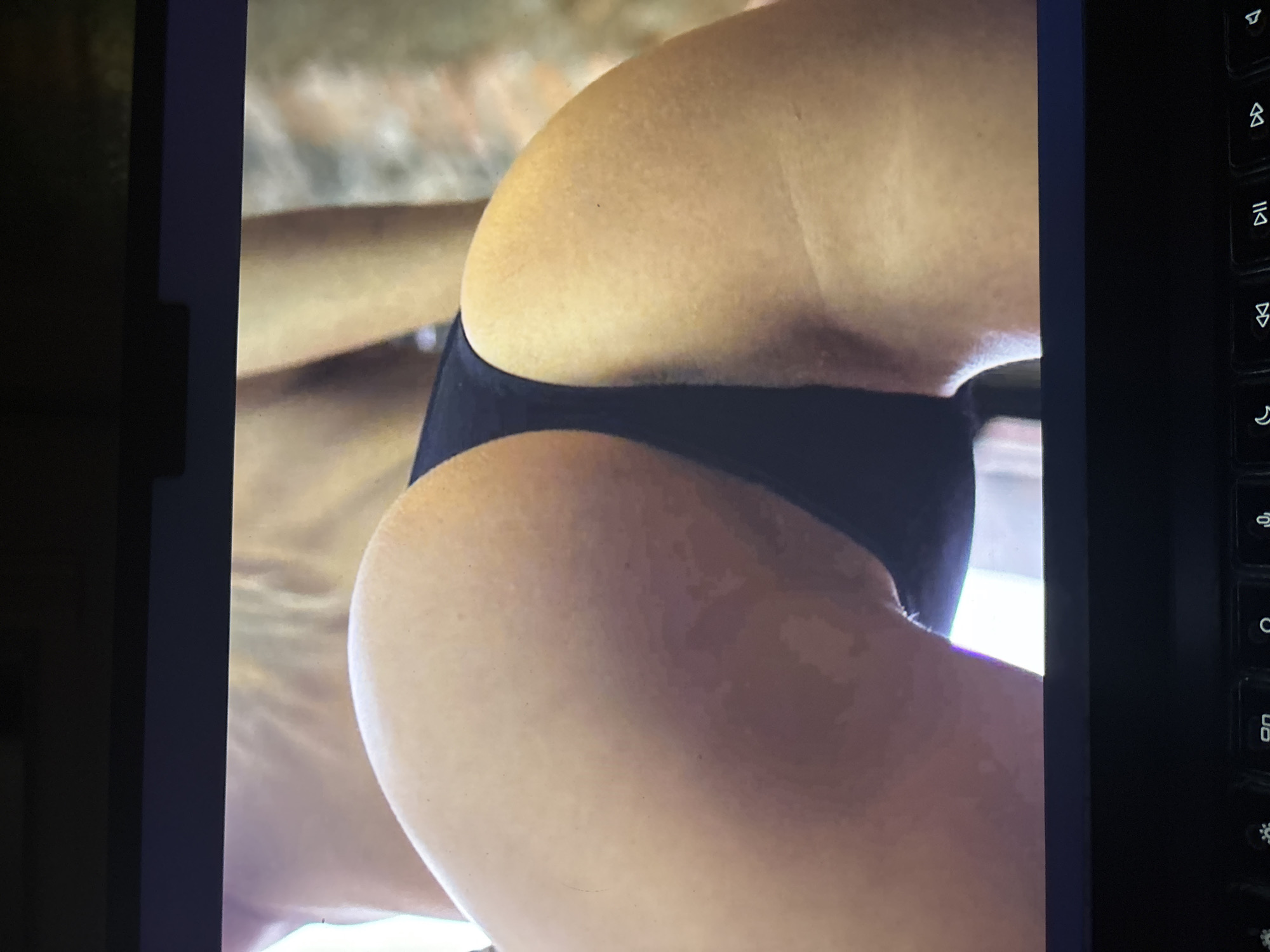 A digital photograph displays a close-up view of a person's lower body, specifically the buttocks and upper thighs, clad in black, form-fitting underwear or bikini bottoms. The skin tone is warm and smooth, with subtle variations in light and shadow highlighting the body's contours. The background within the displayed image is blurred and lighter, suggesting soft, indirect lighting in an undefined indoor or shaded outdoor setting.
The image is presented on a digital screen, with a dark screen bezel visible on both the left and right edges. To the immediate right of the screen, a segment of a black computer keyboard is visible. Several keycaps are discernible, featuring white symbols such as media controls (e.g., play/pause, skip forward, skip backward), a mute icon, and brightness adjustment symbols (sun with up/down arrows), along with a moon symbol that often indicates a sleep or power function. The overall impression is an image of a person viewed on a display, framed by the digital device it's being shown on, likely in an indoor setting in Sofia, Bulgaria.