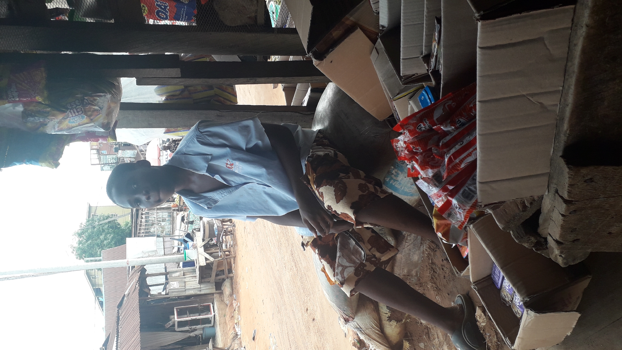 A person wearing a light blue collared shirt and floral patterned shorts sits amongst stacked cardboard boxes and bags of goods inside a makeshift structure. The person appears to be in their teens. The location is Nkpor, Nigeria, as evidenced by the architecture and environment visible through the opening of the structure. Outside, there are buildings with corrugated metal roofs and a dirt road. The area seems to be a commercial or residential area.