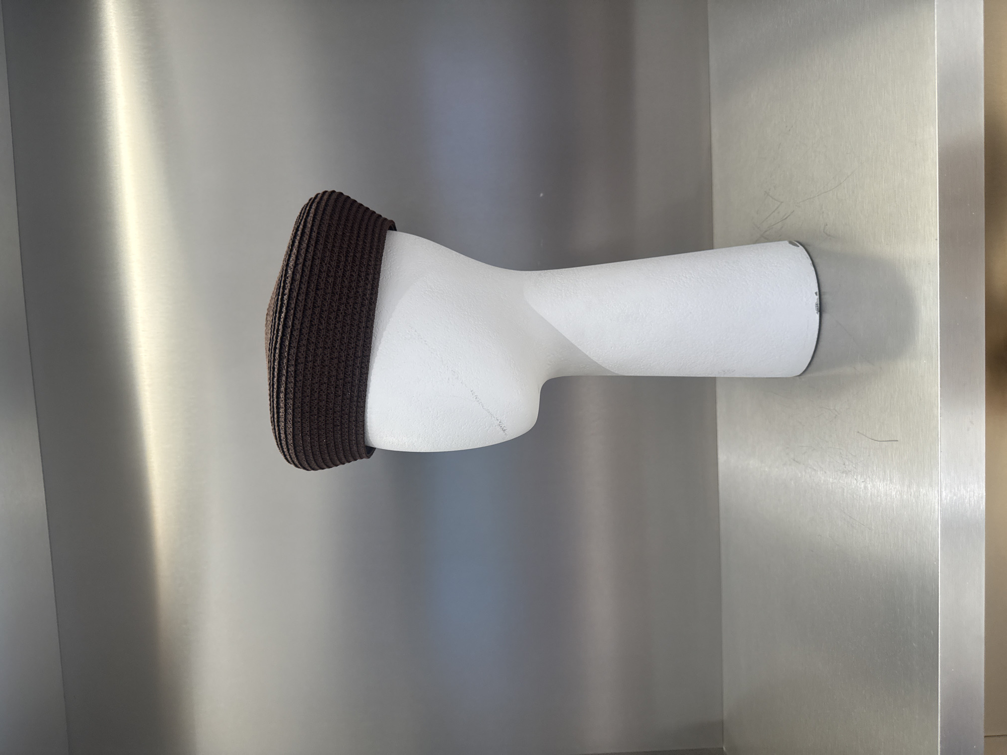 A brown knitted cap is placed on a white mannequin head inside what appears to be a modern or minimalist space, possibly a store or exhibit. The background is a light gray surface, potentially a metal or painted wall, with some variations in texture and reflection. The image was taken in Skokie, Illinois, United States of America.