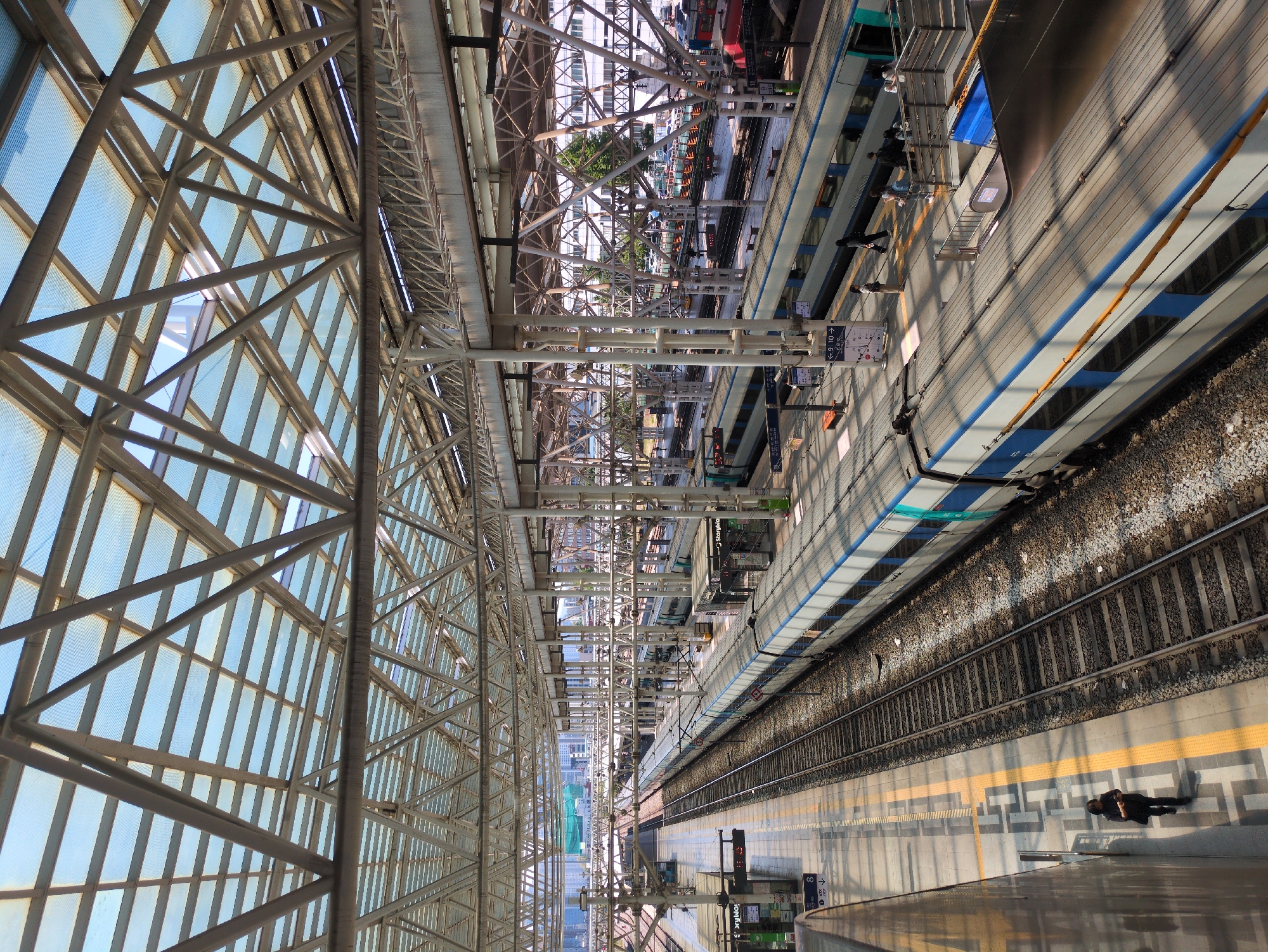 An eye-level shot looking down a Seoul train station platform. On the left, a metal and glass roof arch. A pair of train tracks extends lengthwise. A passenger walks along the platform. A train car sits to the right. Overhead, more steel architecture.