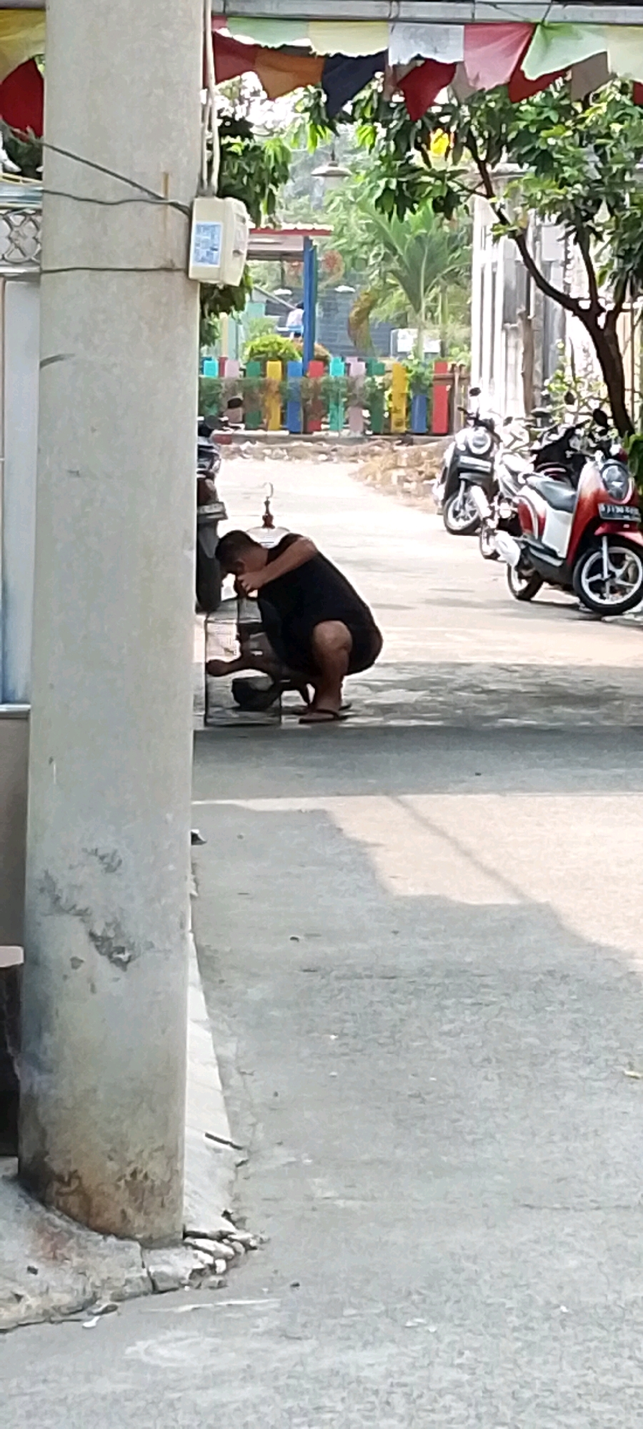 In Satriamekar, Indonesia, a person wearing a black t-shirt and shorts squats next to a motorcycle, appearing to examine something on the ground. The scene takes place outdoors on a paved surface. In the background, colorful decorative elements are visible, along with other motorcycles parked. A utility box is mounted on a concrete pole to the left of the frame.