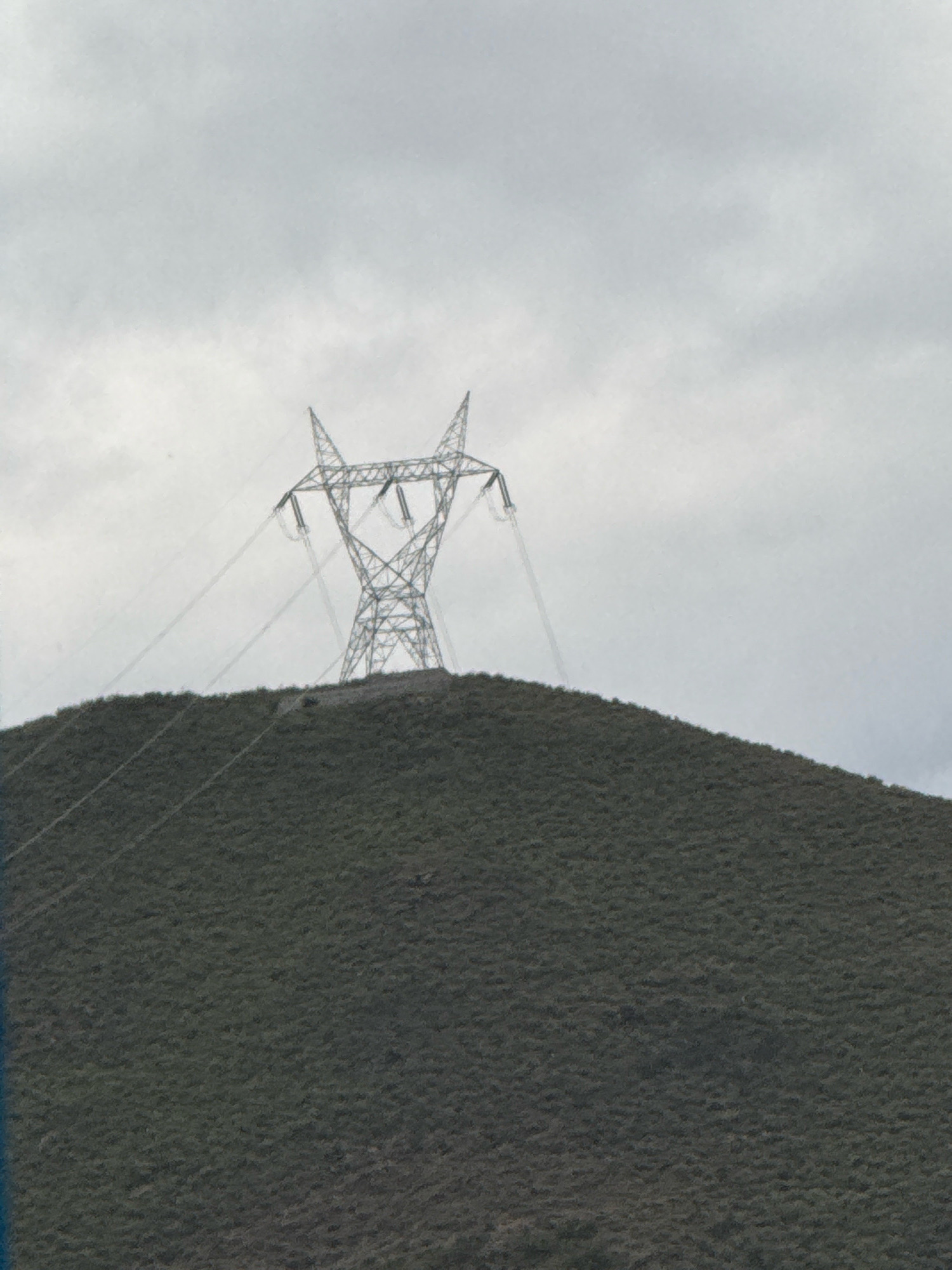 A wide shot shows a large, metal power line tower standing atop a grassy, rolling hill. The sky above is overcast and gray. The tower is the dominant feature in the center of the frame, with its lattice structure clearly visible. Several thick cables extend from the tower and disappear down the side of the hill. The hill itself is covered in low-lying vegetation, creating a textured surface. The scene appears to be in a rural or semi-rural area, with no buildings or other signs of urban development immediately visible. The overall impression is one of a functional piece of infrastructure set against a natural landscape under a somber sky.