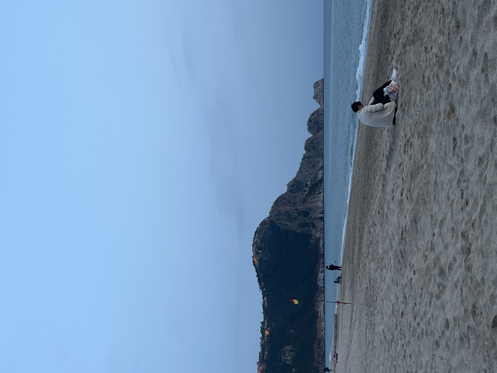 A medium shot of Alanya, Turkey, focuses on the beach. The sky is overcast. A land formation covered in foliage stretches from the middle to the bottom of the image. The ocean meets the sand; white foamy waves are visible. The beach takes up the right side of the image. Two individuals are sitting near the water. Further down the beach, a person walks near two beach chairs, a red flag, and a yellow and red flag. A building, likely a hotel, is visible at the bottom.