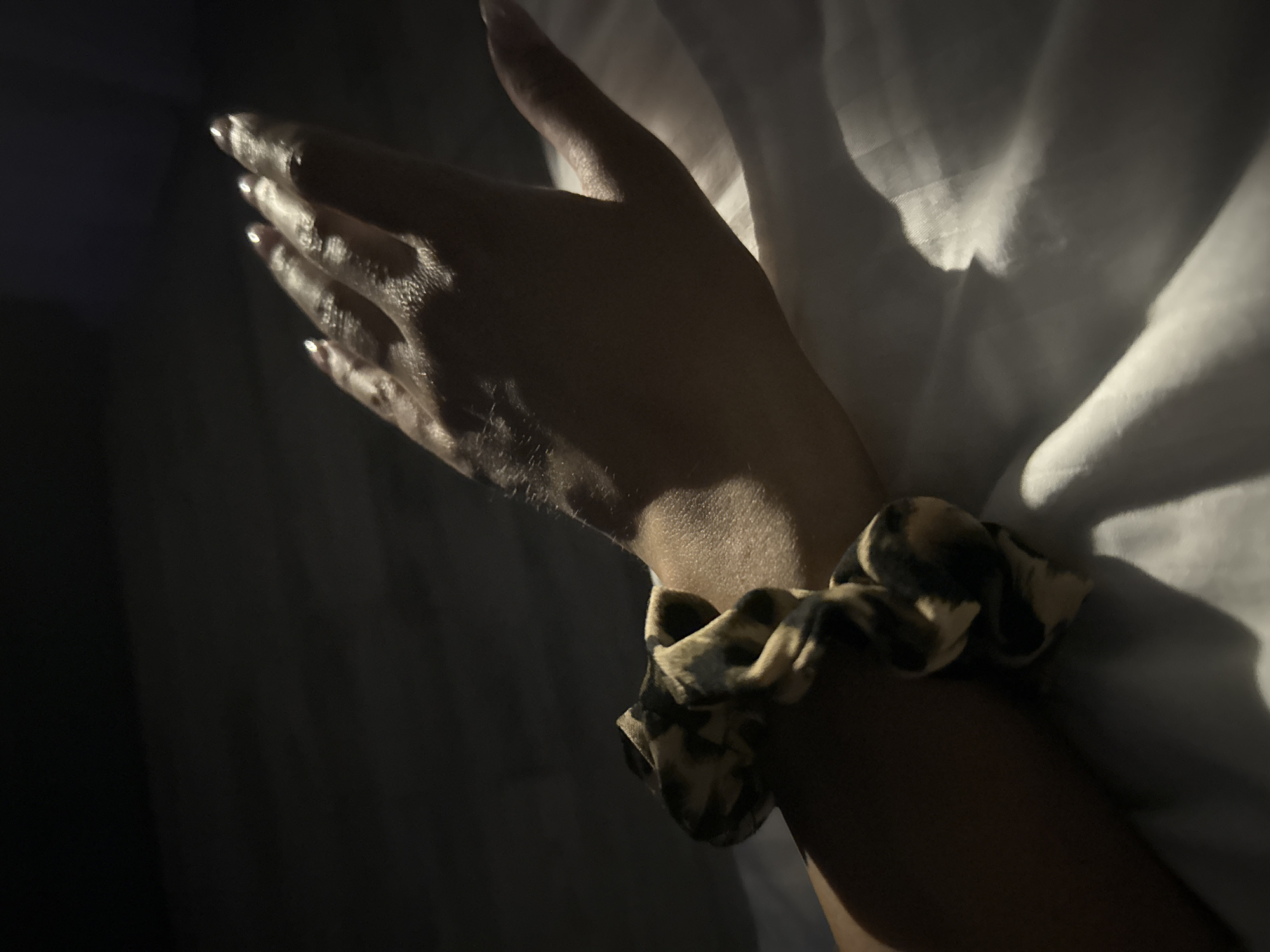 The image shows a close-up of a person's left arm and hand, adorned with a scrunchie. The arm is resting on a white surface, possibly a bedsheet, which is illuminated by a dramatic spotlight. The hand is positioned above the arm, with fingers slightly splayed. The nails appear to be manicured. The scrunchie has a leopard print pattern in shades of beige and black. The background is dark, suggesting low lighting conditions. There is no discernible information about the location in the city of Doma 1096 Km, Russia, or any specific activities taking place beyond the pose of the arm and hand.