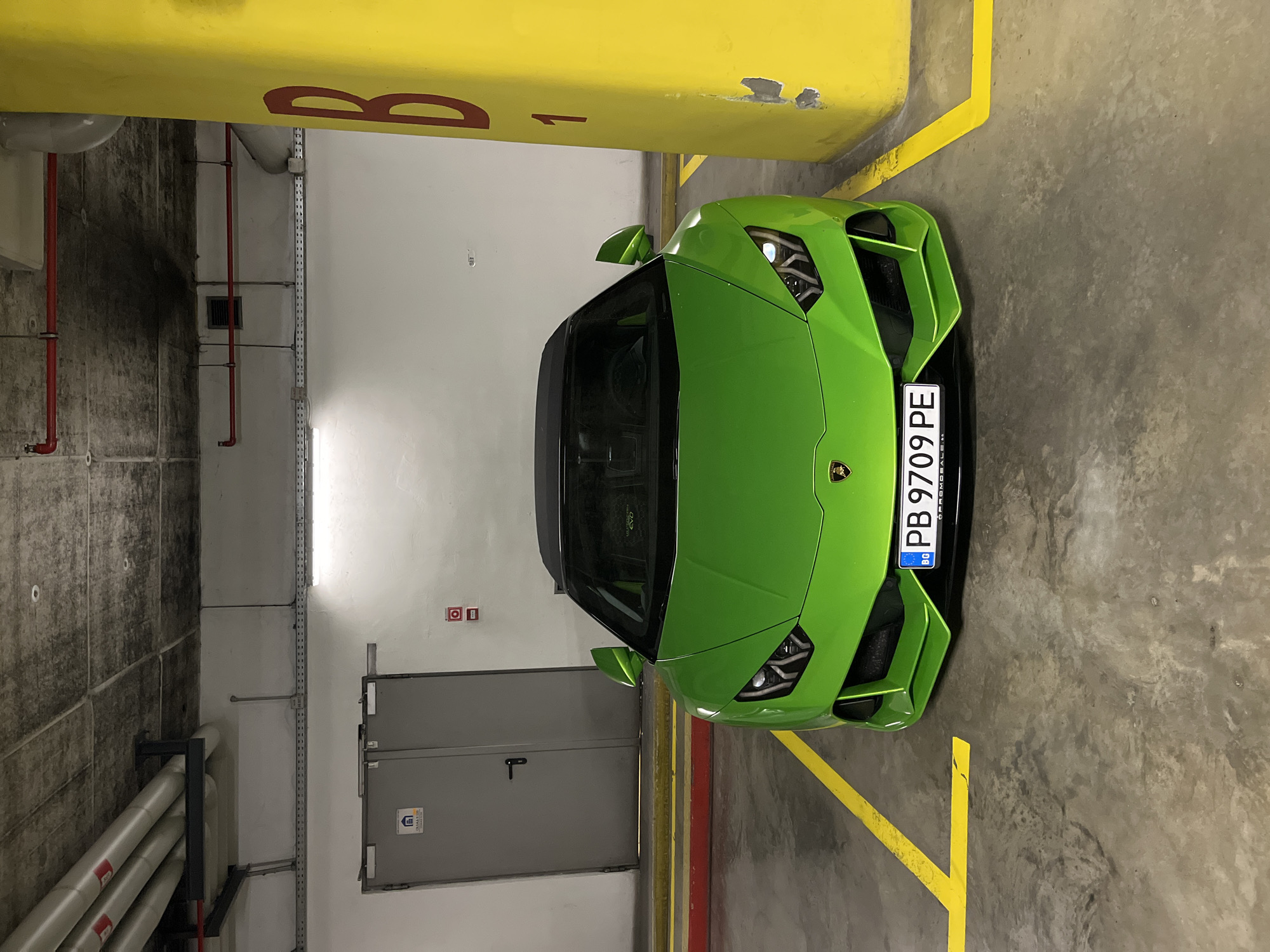 The image shows a green Lamborghini sports car parked in an indoor parking garage. The car has a Bulgarian license plate that reads "PB 9709 PE". The parking space is marked with yellow lines on a gray concrete floor. The garage has yellow-painted support columns, with a "B" designation. A gray utility door and pipes run along the left side of the image. The exact location in the city of Plovdiv, Bulgaria, cannot be determined.