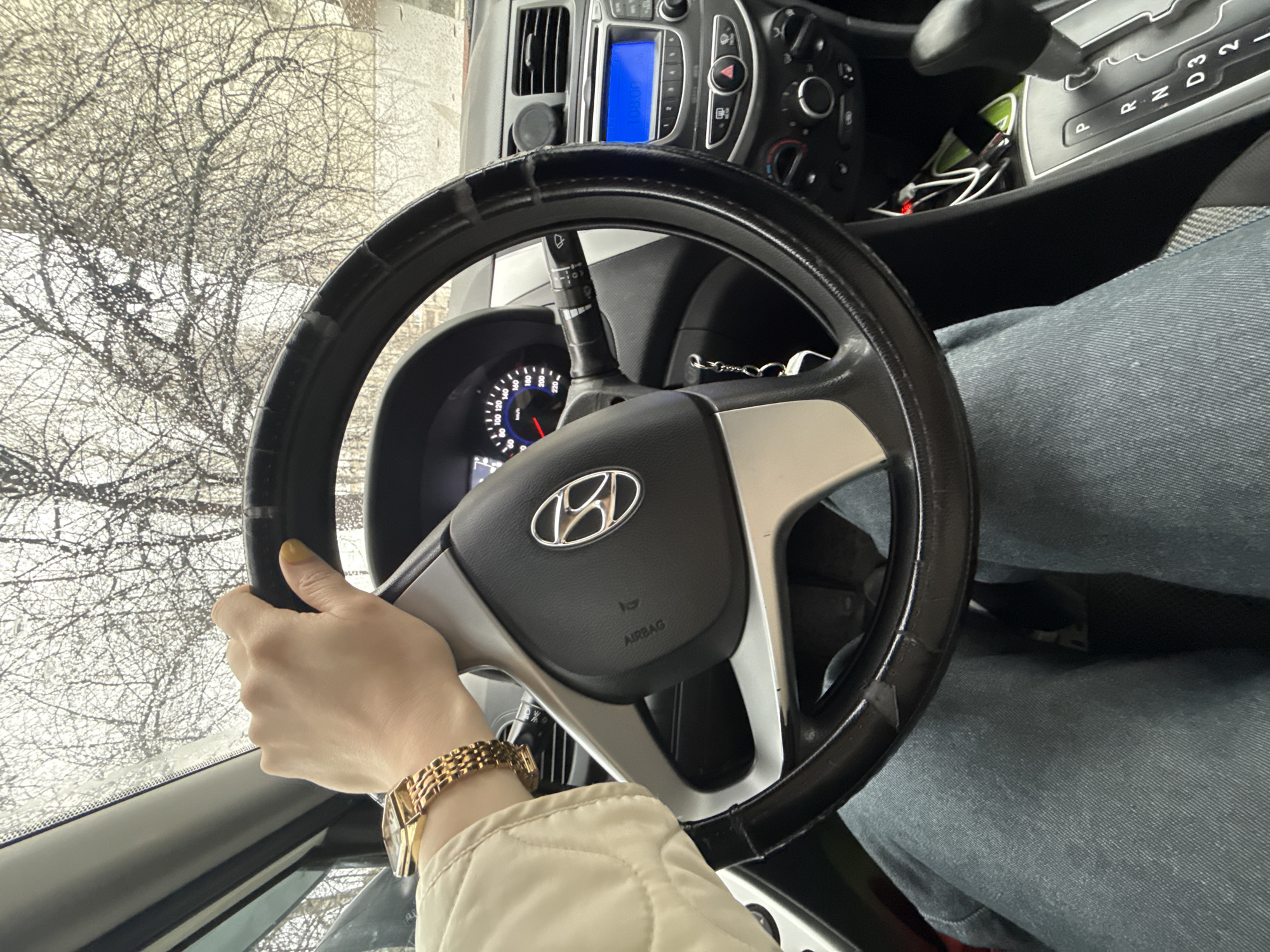 A person is shown behind the wheel of a Hyundai car. The driver is gripping the steering wheel, wearing a gold wristwatch and a light-colored jacket. Outside the car window, a tree without leaves can be seen. The location is unknown and there is no evidence of the image being located in the city of Chaikovsky, Russia.