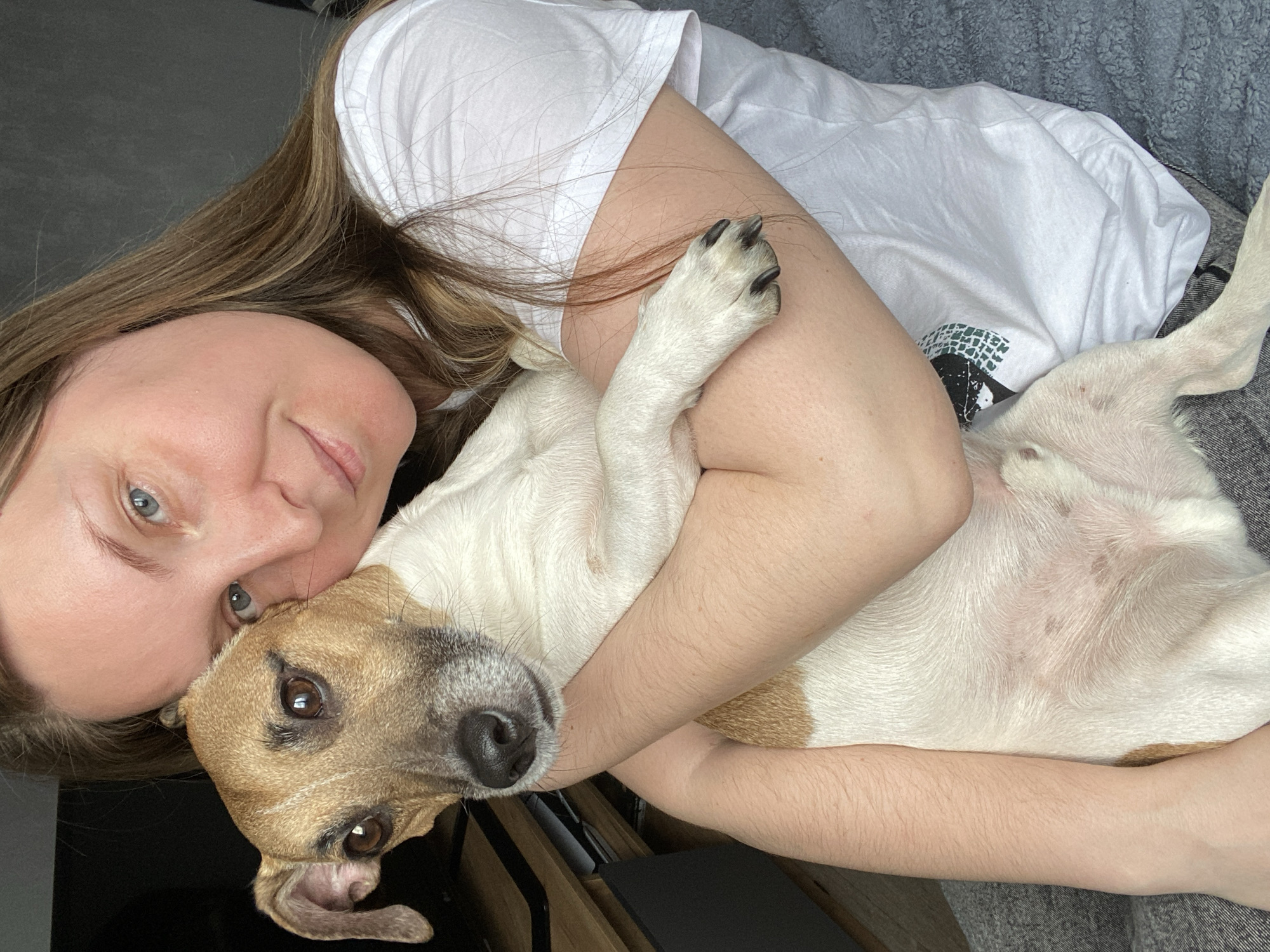 An indoor, overhead shot captures a woman embracing her dog. The woman, with light skin, light brown hair, and blue-grey eyes, lies on her side, her head tilted, looking towards the viewer with a gentle expression. She is wearing a white short-sleeved t-shirt with a small, indistinct dark green and black graphic on the left chest area. Her right arm is wrapped around a small to medium-sized dog, holding it close to her body.
The dog has white fur with distinctive brown patches on its head, particularly around its ears and eyes, and a dark snout. It is snuggled against the woman, appearing relaxed, with its paws resting on her arm. The scene conveys a strong sense of affection and companionship. The lighting is soft and natural, suggesting daytime. The background includes a dark gray surface and a lighter gray textured fabric, possibly a blanket or bedding, indicating a cozy, personal setting within a home in Noviy, Russia. No readable text is visible.