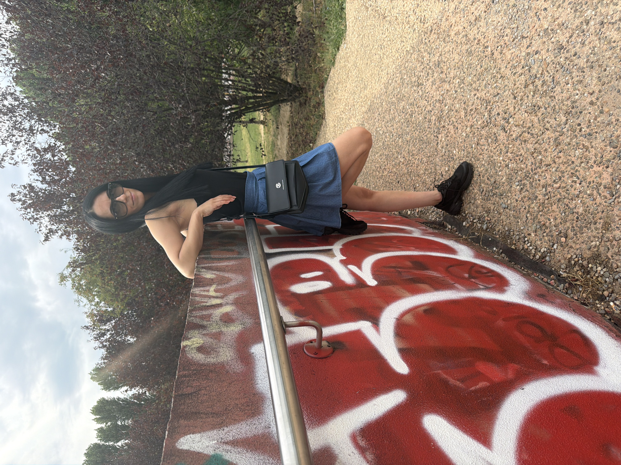 A woman with long, dark hair and sunglasses leans against a graffiti-covered wall in an outdoor urban setting. She is wearing a black tank top, a denim skirt, and black athletic shoes. A black crossbody bag is slung over her shoulder. The wall, painted red with white and yellow graffiti, features a silver handrail. The ground is a textured, beige pathway. In the background, trees with dark red and green foliage are visible under a cloudy sky. The scene appears to be in a park or public space.