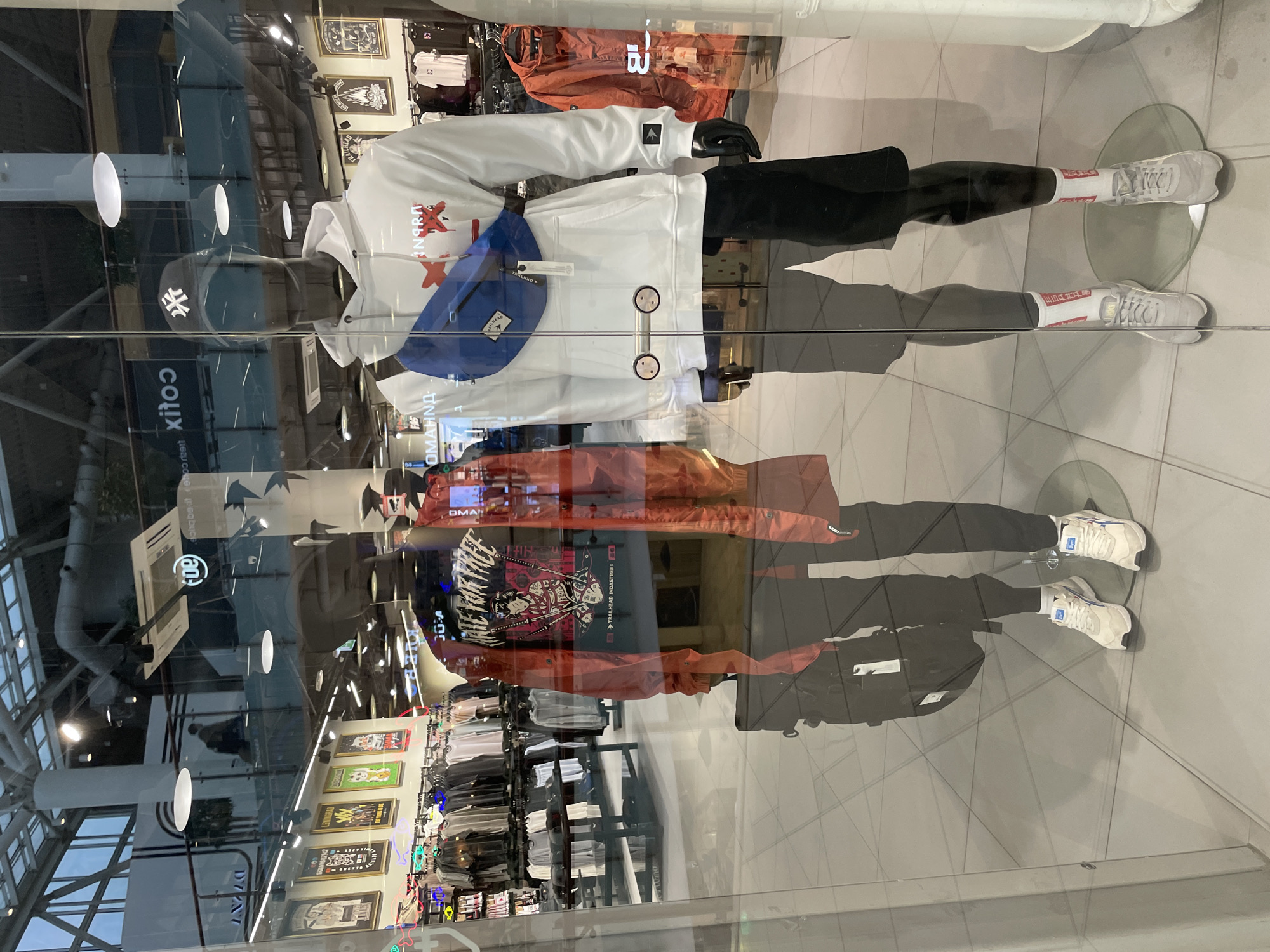 A display window in a store in Khimki, Russia, reflects an interior view of the store as well as a part of the ceiling of the building housing the store. The display features two mannequins. The top mannequin wears a white hoodie with red designs, a blue waist bag, black shorts, white socks with red details, and white sneakers. The second mannequin is clad in an orange jacket, a black shirt with a white graphic design, black pants, white socks with red details, and white sneakers. Clothing racks filled with items are visible inside the store. Signage and framed art line the back wall. The word "xitoo" is visible on a sign.