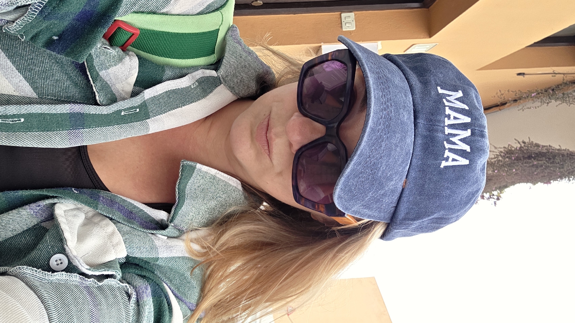 A medium shot shows a woman wearing a blue baseball cap with "MAMA" embroidered in white, large dark-rimmed sunglasses, and an open green, white, and purple plaid shirt over a black top. A green and white striped strap with a red buckle crosses her chest, suggesting a bag or backpack. Her blond hair is visible on the right side of her face. The background consists of an orange building to the left and a light-colored wall to the right, with some greenery hanging from above. The scene appears to be outdoors, possibly on a street or walkway in Guía de Isora, Spain, indicated by the architectural style.