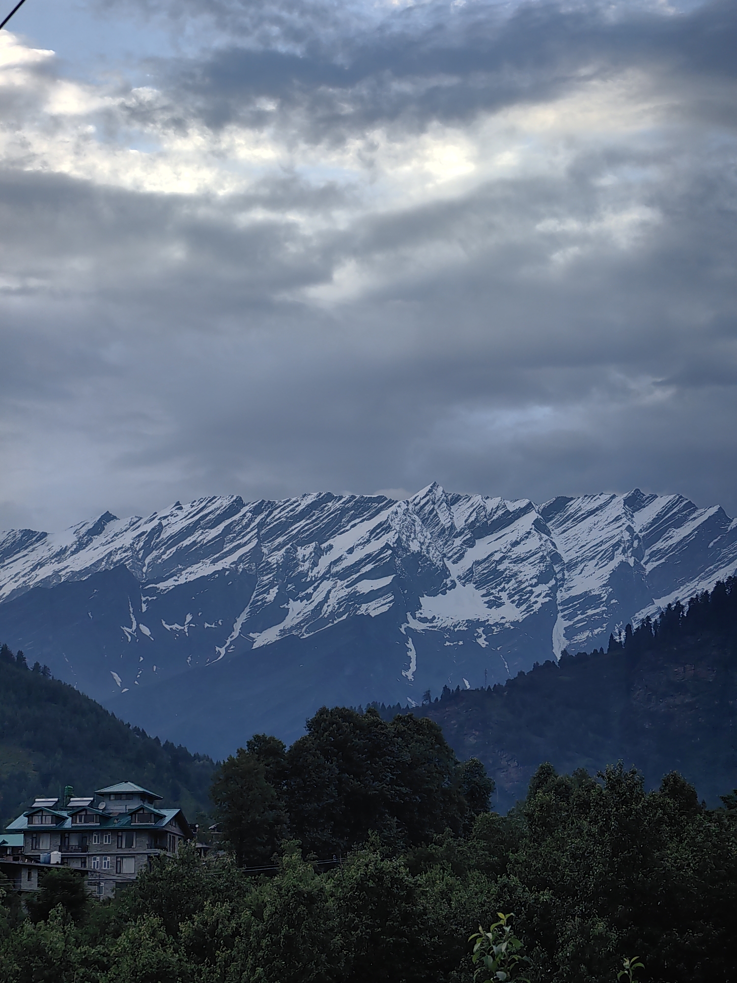 This image captures a serene mountain landscape in Kulang, India, likely during the late afternoon or early evening given the soft, diffused light. The sky is filled with dramatic, heavy clouds, suggesting an overcast or approaching storm, yet there are patches of brighter sky peeking through, hinting at the sun's presence.
The focal point of the scene is a majestic mountain range with snow-capped peaks. The snow appears to be streaked, perhaps from recent snowfall or wind erosion, creating a textured and rugged appearance against the darker slopes. The mountains are partially shrouded in shadow, adding to their imposing presence.
In the foreground, a lush, dense forest blankets the lower slopes of the mountains. Amidst the greenery, a large, multi-story building with a distinct green roof stands out. The architecture suggests a hotel or resort, blending with the natural surroundings yet clearly man-made. The building has many windows and balconies, and appears to be constructed with stone or concrete. The trees in the foreground are deep green, creating a stark contrast with the white snow of the distant mountains.
No people or specific activities are visible. The overall mood is one of natural grandeur and tranquility, with a hint of dramatic weather. The location cues, such as the architecture and the type of mountains, strongly suggest the Himalayan region. No text is visible in the image.