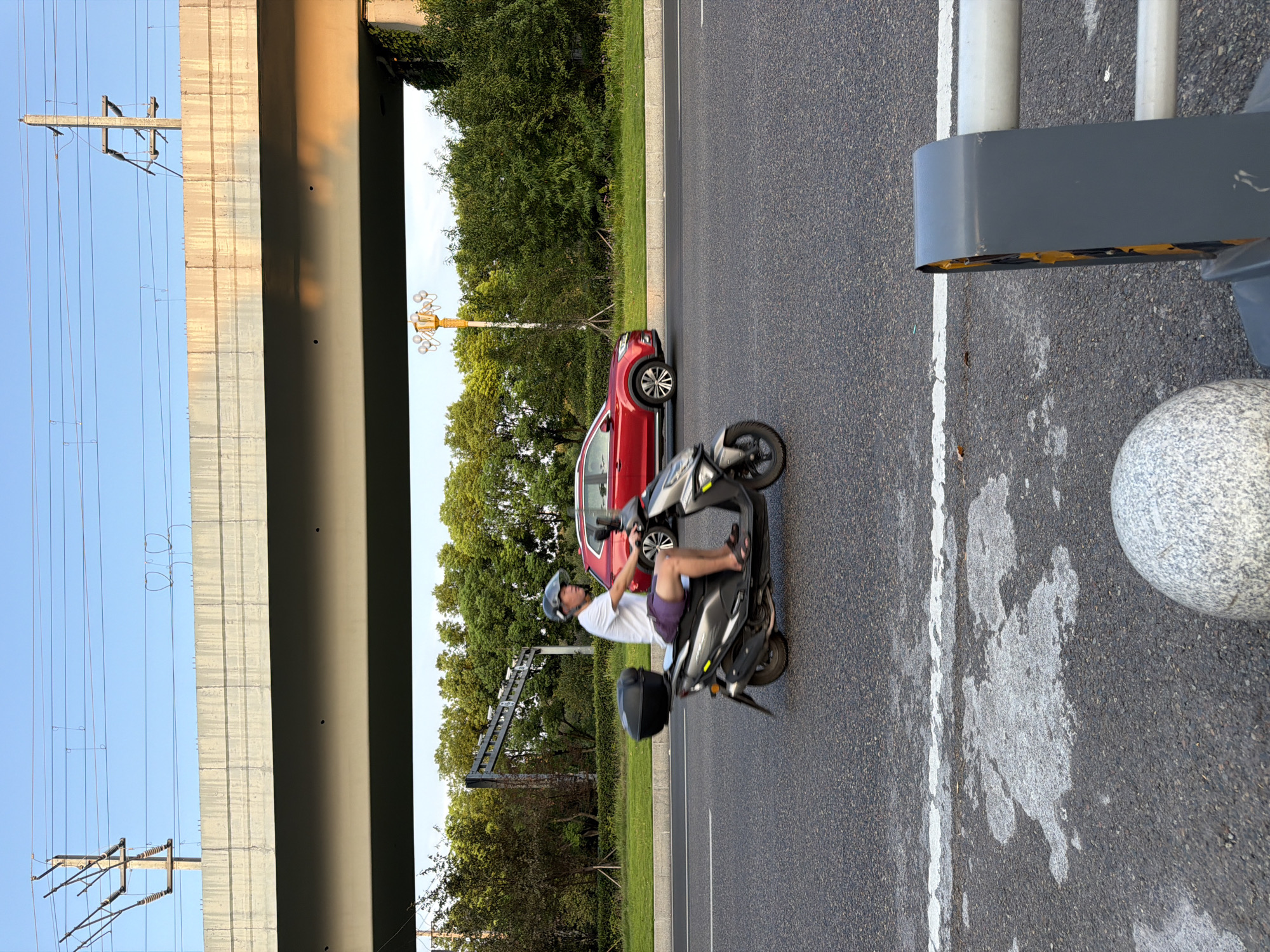 An outdoor, eye-level, medium shot shows a man riding a scooter on a paved road under an elevated highway structure. The man, wearing a helmet and a white t-shirt with purple shorts, is in the foreground and is captured in motion. Behind him, a red SUV is also moving on the road. The background consists of lush green trees and a clear blue sky. Overhead, a concrete overpass with electrical wires and poles is visible. The lighting suggests it is either early morning or late afternoon. The foreground also includes a stone bollard and a section of a road barrier.