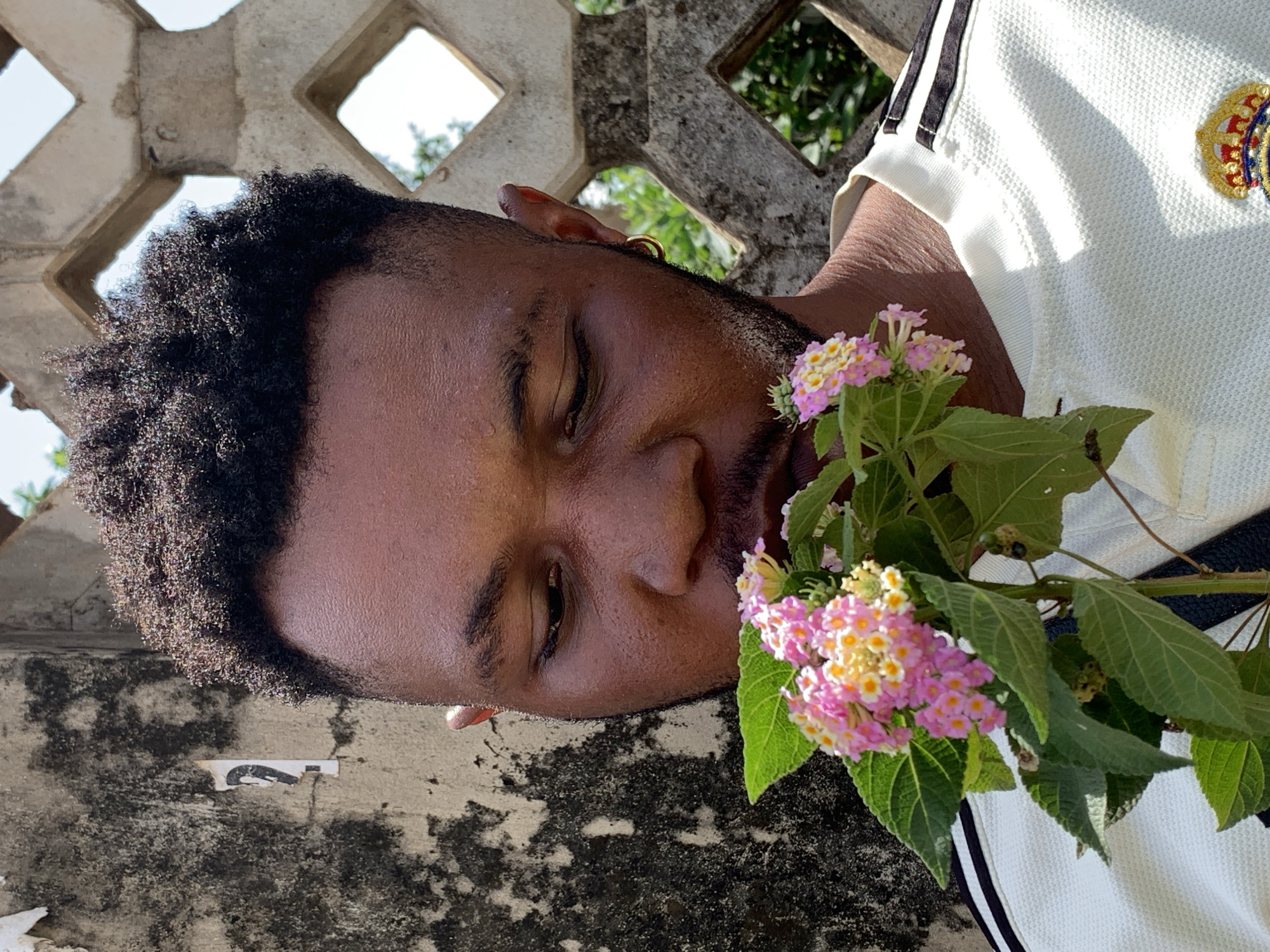 The image shows a man with dark hair and skin lying down on a concrete surface next to a partially visible concrete block wall with diamond-shaped openings. He is wearing a light-colored shirt with black stripes on the shoulder. A sprig of pink and yellow flowers with green leaves is positioned near his face. The background includes some greenery visible through the openings in the wall. There are no explicit indicators that the location is Ikorodu, Nigeria.