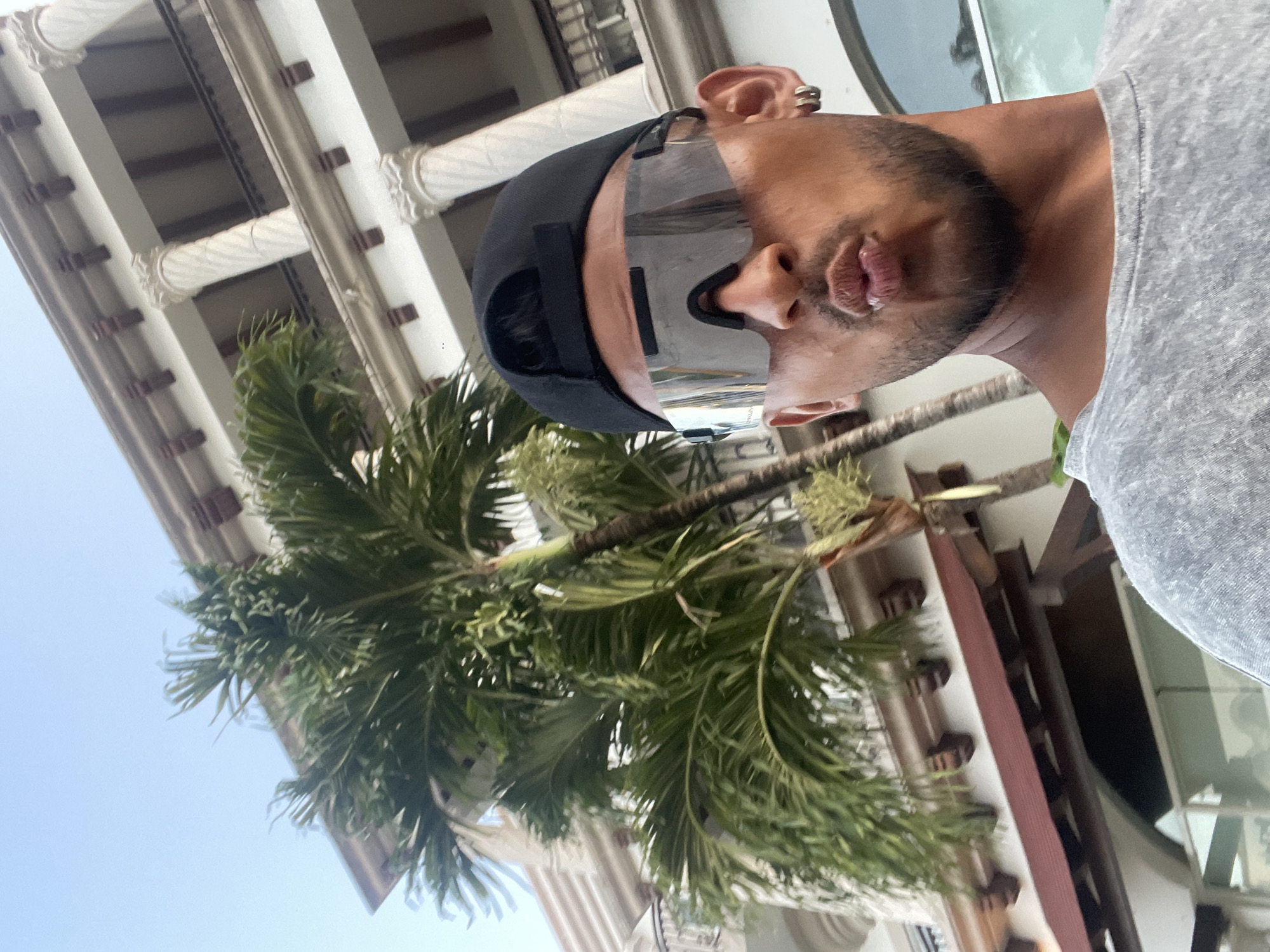 A medium shot shows a person wearing a gray shirt, black cap, and silver futuristic sunglasses, with an ear piercing, positioned against a backdrop of a tall, ornate building with white columns and a palm tree. The photo is taken from a low angle, looking up at the person and the building. The location is identifiable as Playa del Carmen, Mexico, due to the architectural style and the presence of a palm tree.