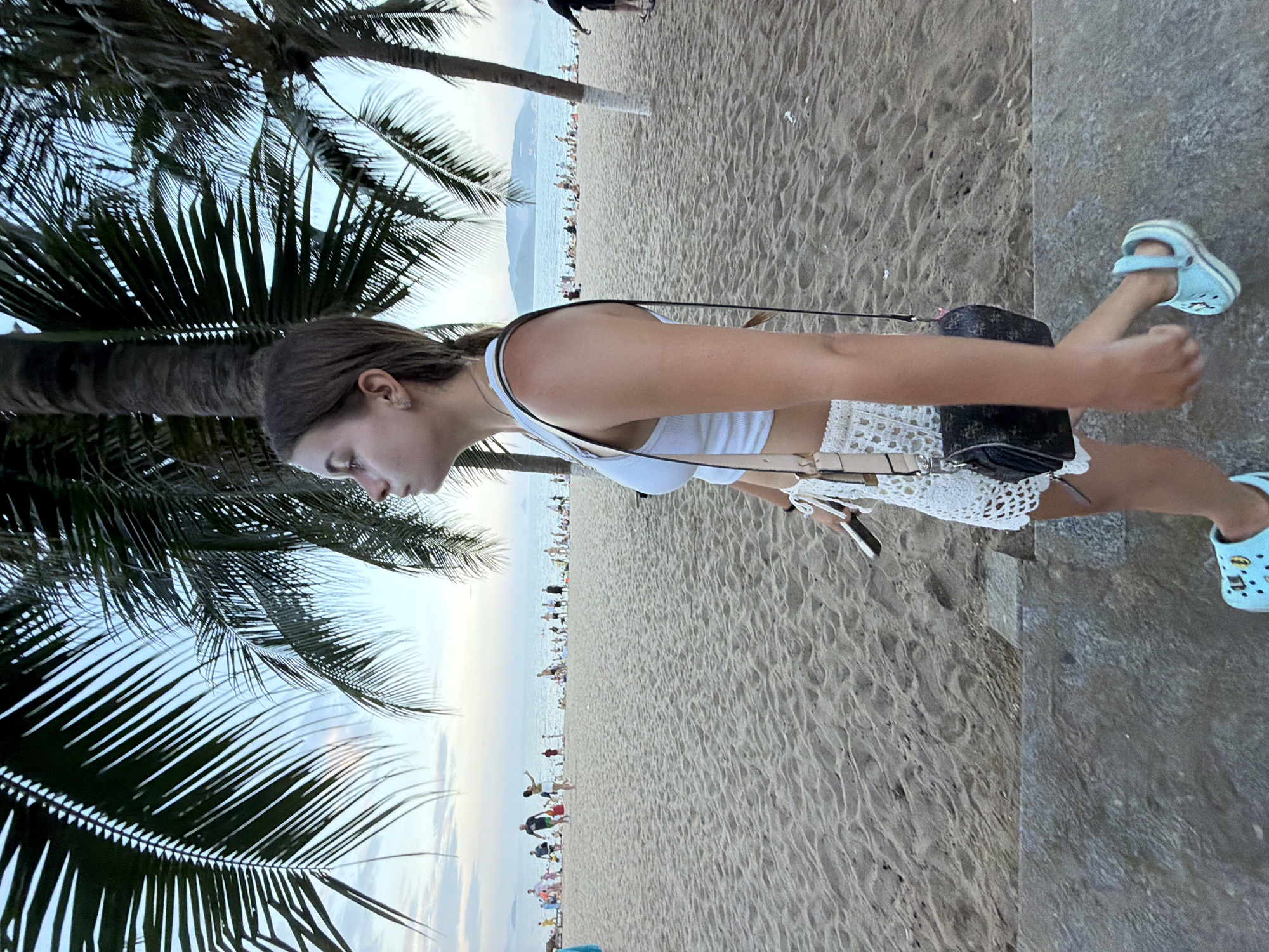 The image captures a young woman walking on a paved path adjacent to a sandy beach in Nha Trang, Vietnam. The woman, seen in profile, wears a white crop top and white crochet shorts, with a small patterned crossbody bag slung over her shoulder. She also has light blue crocs on her feet. In the background, the beach is populated with numerous individuals, some relaxing, others walking, and a few playing in the shallow water. Palm trees frame the scene, their fronds extending into a sky with scattered clouds. The sea stretches to the horizon, with a distant island visible. The lighting suggests it is late afternoon or early evening.