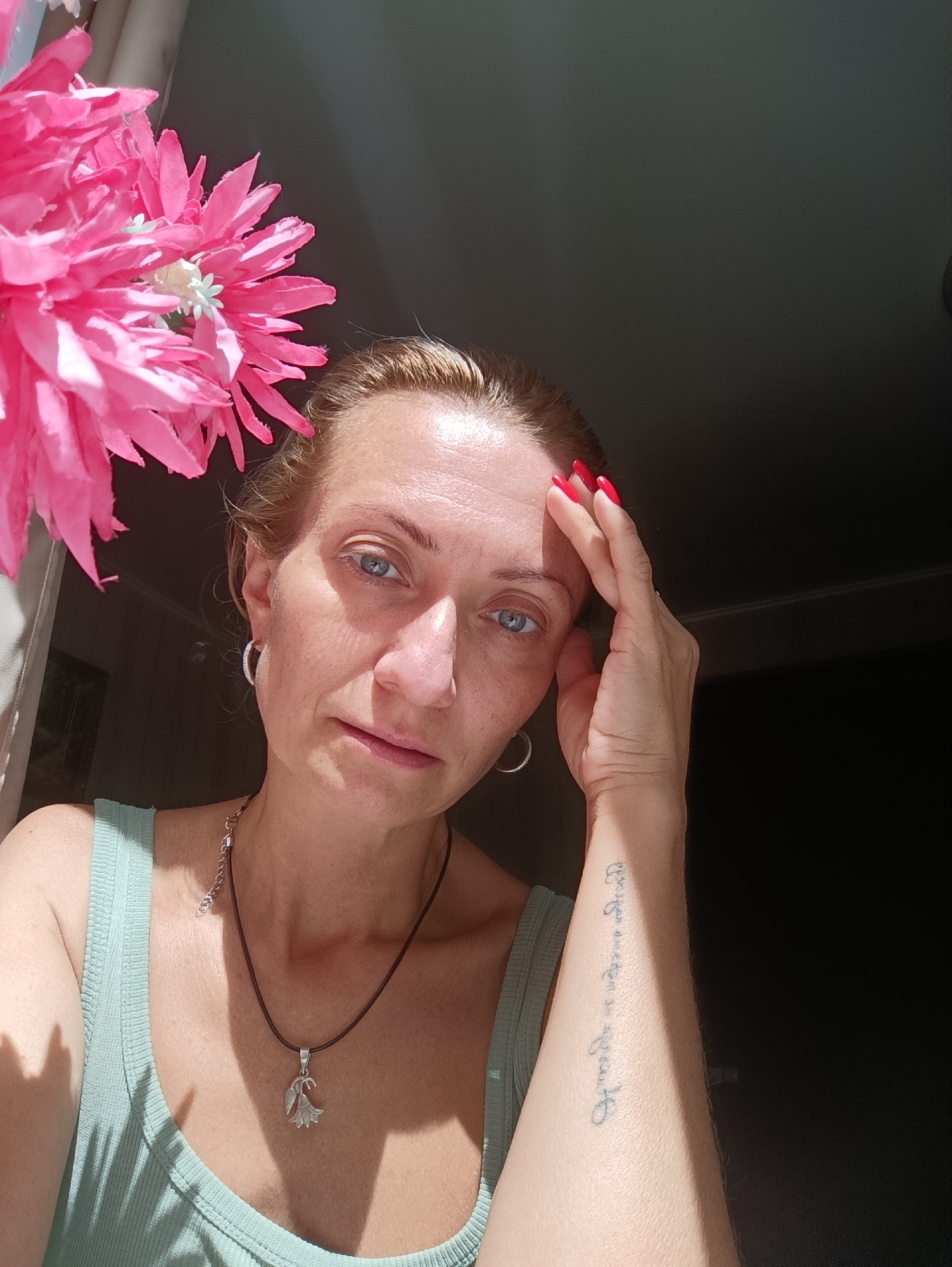 A woman with blonde hair and blue eyes is looking at the camera. She is wearing a light green tank top, a silver necklace with a flower pendant, and hoop earrings. Her right hand is touching her forehead, and her fingernails are painted red. There is a tattoo on her left forearm with vertical writing. A large pink floral decoration is in the upper left corner of the image, casting a shadow. The background is dark. The image appears to have been taken indoors, near a window, as indicated by the shadows and the light source. The overall scene suggests a casual, indoor portrait. The prompt specifies the location as Chaikovsky, Russia, but there are no visual elements in the image that can confirm or deny this.