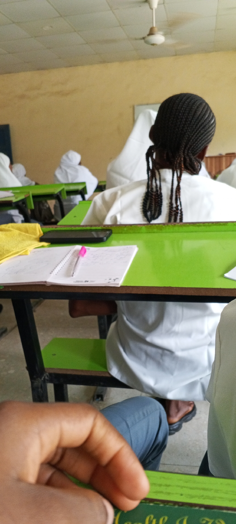 The image depicts a classroom setting. Students in white uniforms are seated at green desks. A notebook, pen, and smartphone are on a desk in the foreground. A person's hand and leg are visible in the lower part of the image. The location is unknown.