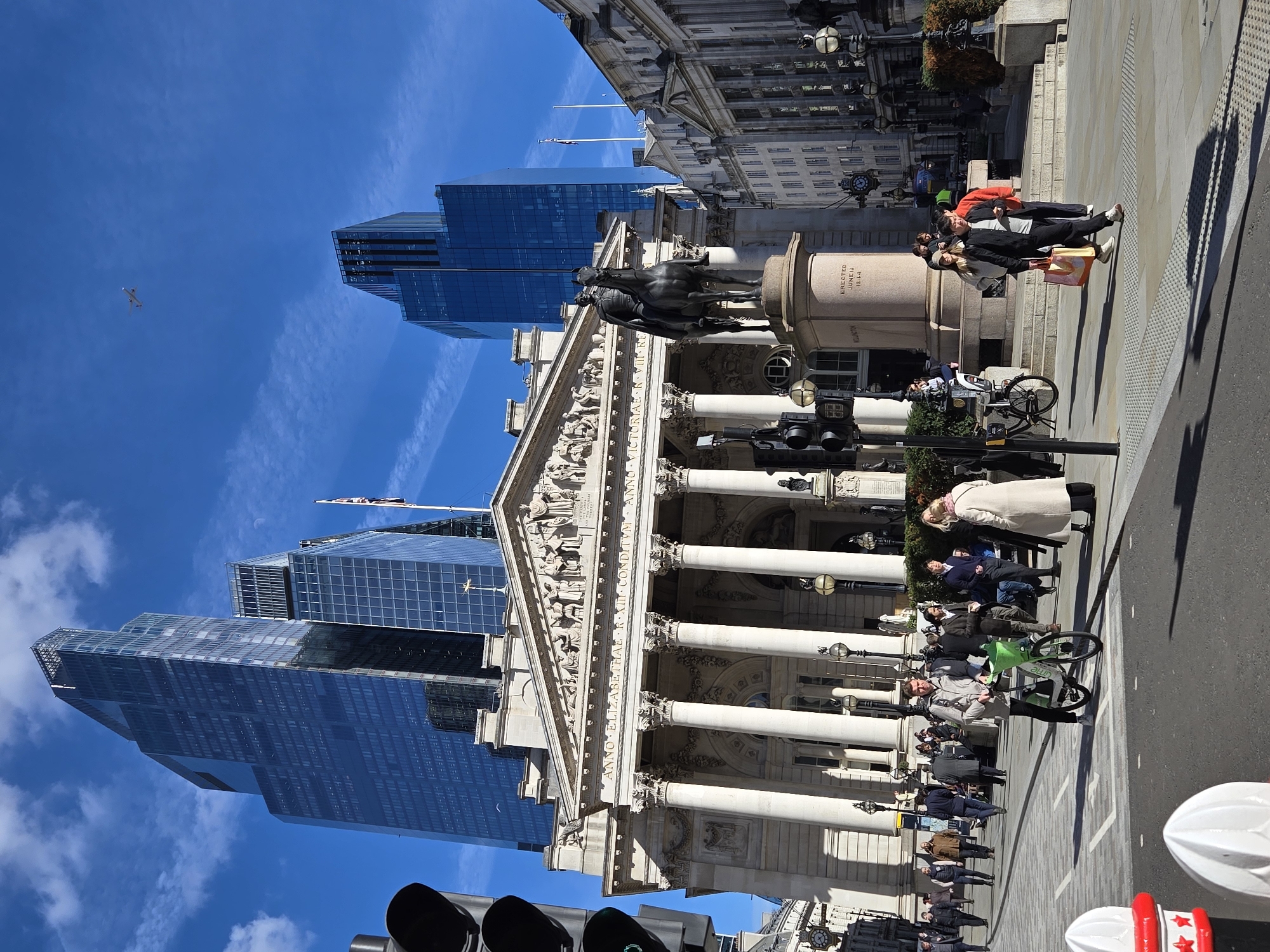 This image captures a vibrant daytime scene at Bank Junction in the City of London, United Kingdom. The Royal Exchange, a grand Neoclassical building with a prominent portico featuring eight Corinthian columns and an ornate sculptural pediment, stands centrally in the background. Inscriptions "ANNO ELIZABETHAE R. XIII CONDITUM" and "ANNO VICTORIAE R. VII. REDIFICATUM" are visible on its facade. To the right of the Royal Exchange's main entrance, an equestrian bronze statue of the Duke of Wellington is positioned on a stone plinth inscribed "ERECTED JUNE 1844".
Dominating the upper background are several modern glass skyscrapers, notably 22 Bishopsgate with its distinctive stepped profile, reflecting the bright blue sky. Scattered white cumulus clouds are present, and a small, light-colored aircraft is visible in the upper right sky. A faint crescent moon is also discernable near the top of one skyscraper.
At ground level, the scene is bustling with approximately 30-40 pedestrians. A man in a light grey jacket and dark trousers stands on the paved area in the left foreground, pushing a green Lime bicycle. Further right, another man in a black jacket walks carrying orange and yellow shopping bags. A woman in a cream-colored coat stands conversing to the right. People are observed walking on the patterned pavements and crossing the asphalt road. Bright sunlight illuminates the scene, casting distinct, elongated shadows of individuals and objects onto the ground. Street furniture includes black traffic light poles on the left and partially visible red-and-white bollards in the immediate foreground. Older, ornate stone buildings with multiple windows line the street to the right of the statue, one flying a Union Flag.