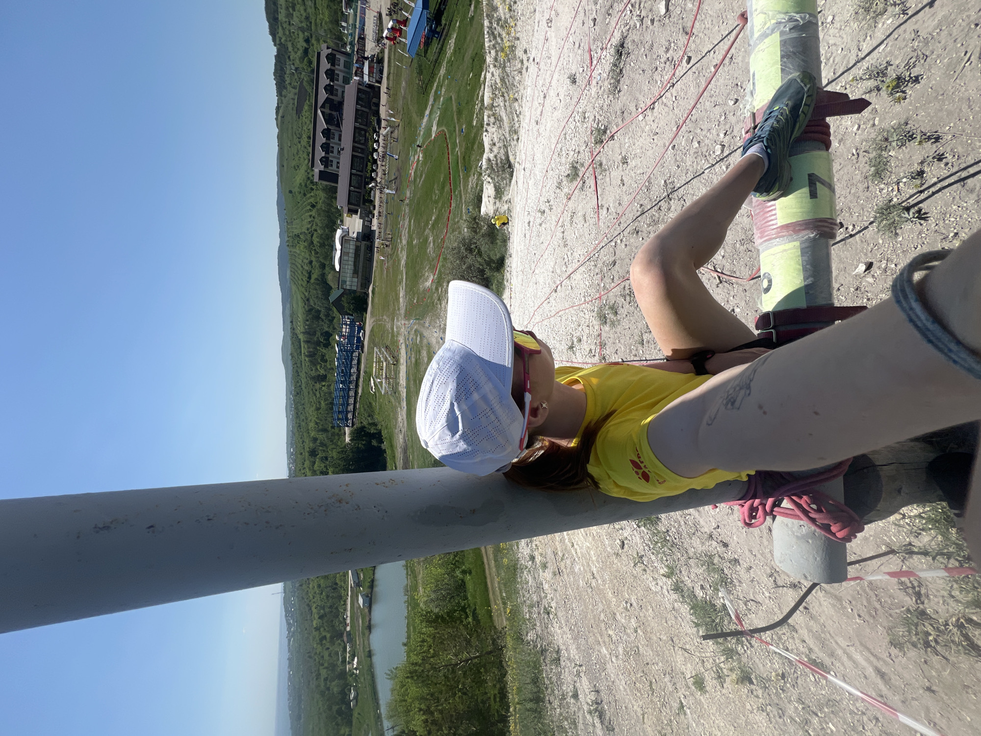 A person wearing a yellow shirt and a white cap is perched high on a structure composed of poles and ropes, likely a climbing or adventure course. The person is secured with red ropes, and their feet rest on a green and yellow pole marked with the number "7". The background features a view of Khvalynsk, Russia, with buildings, trees, and water visible in the distance. The sky is clear and blue, suggesting a sunny day.