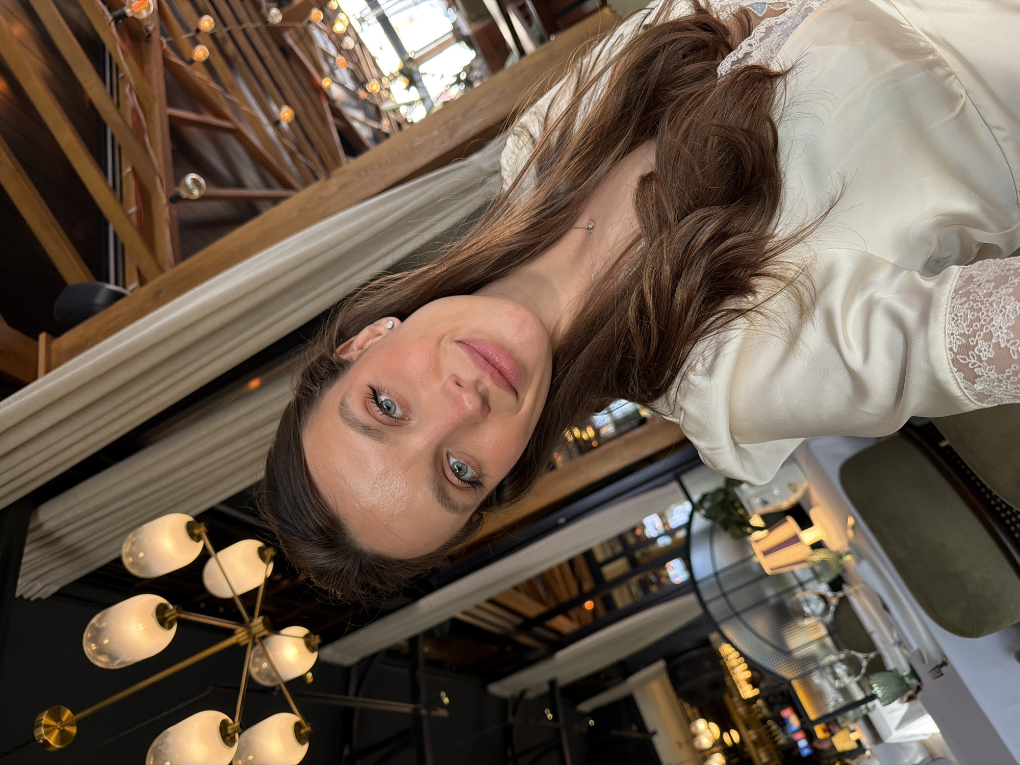 The image displays a slightly angled selfie of a young adult female taken inside an upscale restaurant in Budva, Montenegro. The female subject, positioned centrally, has long, wavy brown hair, blue-green eyes, and a subtle smile directed at the camera. She is wearing a cream-colored, satin-like garment with lace detailing visible on the neckline and sleeves. A small silver stud earring is present in her right ear, and a delicate silver necklace with a small pendant adorns her neck.
The restaurant's interior is visible in the background. To the upper left, a modern brass chandelier featuring six frosted glass globe lights is suspended from a dark ceiling. To the upper right and extending into the middle ground, an exposed wooden beamed ceiling structure is strung with multiple strands of warm-toned, possibly Edison-style, string lights. Dining tables set with white tablecloths, white plates, and green-tinted stemware are partially visible in the lower left. Adjacent to these tables are olive-green upholstered chairs. A small table lamp with a striped, conical shade stands on one of the tables. In the deeper background, dark-framed windows or partitions, light-colored drapes, and a dark, possibly metal, decorative screen or fan are present. A dark speaker is mounted near the wooden beams in the upper right. The environment suggests a sophisticated dining or lounge area.