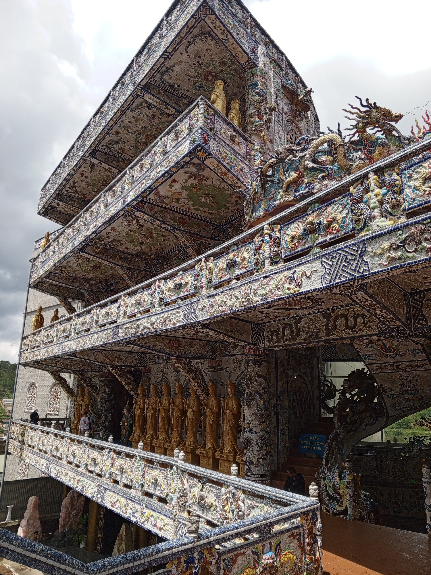 This is an external, low-angle, eye-level shot of the Linh Phuoc Pagoda in Xuan Thanh, Vietnam. The pagoda is a multi-tiered structure adorned with intricate mosaics of ceramic and glass. Several levels of balconies are visible, each decorated with detailed patterns and featuring gold-colored statues of Buddhist figures. A dragon sculpture is prominently displayed on one of the upper balconies. A winding staircase, also decorated with mosaics, leads upwards on the right side of the image. A few individuals are scattered throughout the scene, some walking on the stairs and balconies, while others are standing. The sky above is overcast with clouds. The environment appears to be a tourist attraction, with a sign in Vietnamese visible near the staircase.