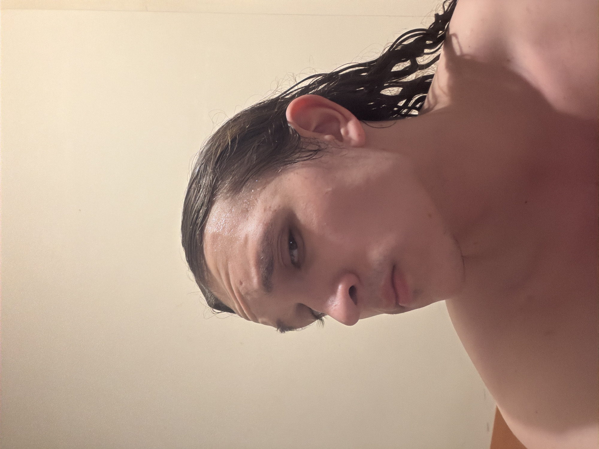 A rotated close-up portrait features a young person, likely male, with long, dark, wet hair. The subject's head is tilted, showing part of their face including one eye, their nose, and mouth, with a neutral or slightly contemplative expression. Their skin appears fair, with a slight sheen on the forehead suggesting wetness or sweat. The upper body, including a bare shoulder and chest area with some redness, is visible, implying they are shirtless or wearing minimal clothing, possibly after a shower or swimming. The background is a plain, light-colored (off-white or beige) wall, indicating an indoor setting. The lighting is soft and even. There is no visible text. The location context for this media is Plovdiv, Bulgaria.