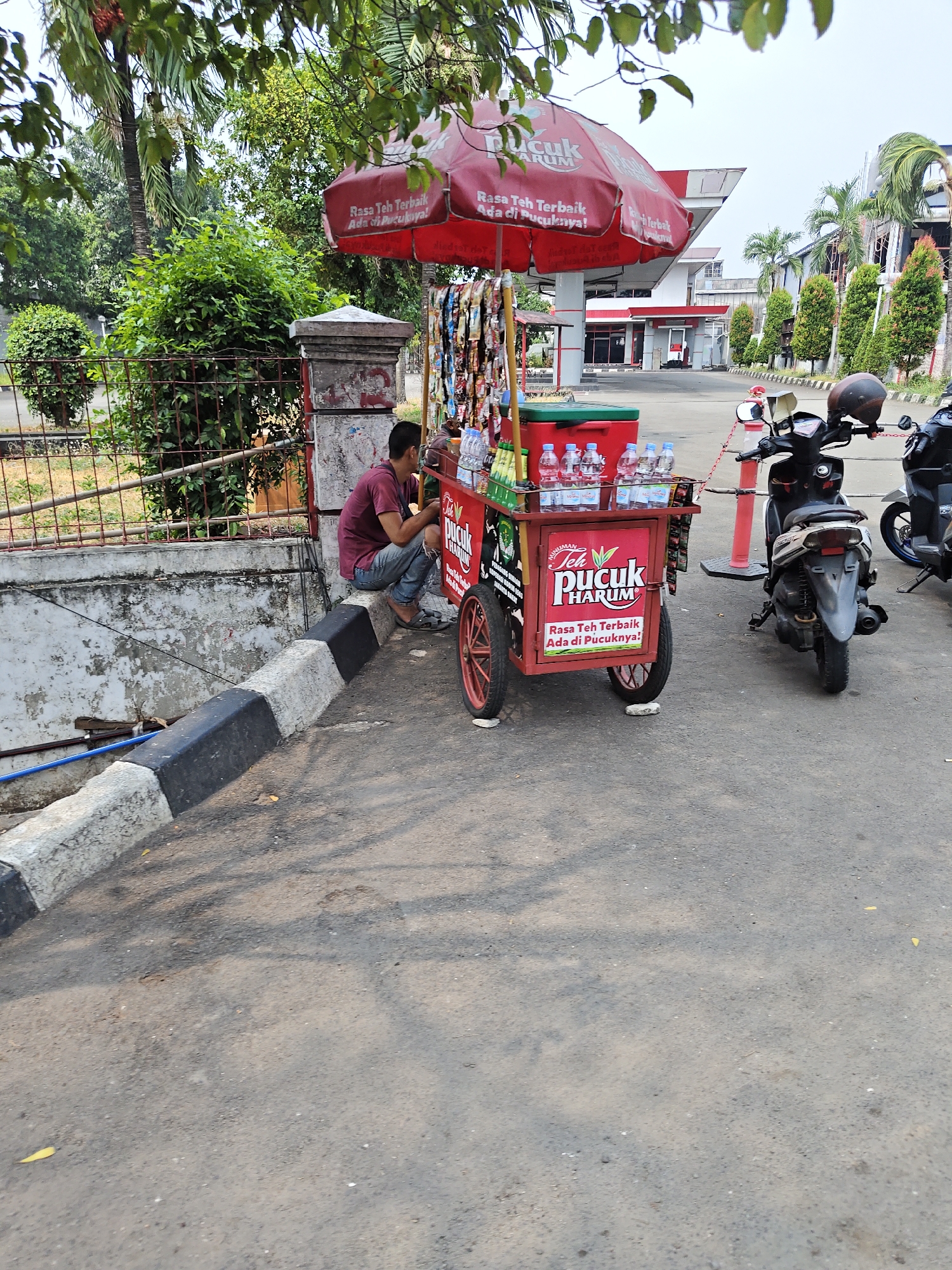 A man in a maroon t-shirt and blue jeans sits on a concrete ledge next to a red and green beverage cart. The cart is topped with a red umbrella advertising "Teh Pucuk Harum" tea. The cart displays bottles of water and tea, as well as other assorted goods. Behind the cart, two motorcycles are parked, one with a helmet on the handlebars. The scene is set at a gas station, indicated by the presence of a gas pump and station building in the background. Trees and foliage are visible around the location. The man appears to be resting or waiting.