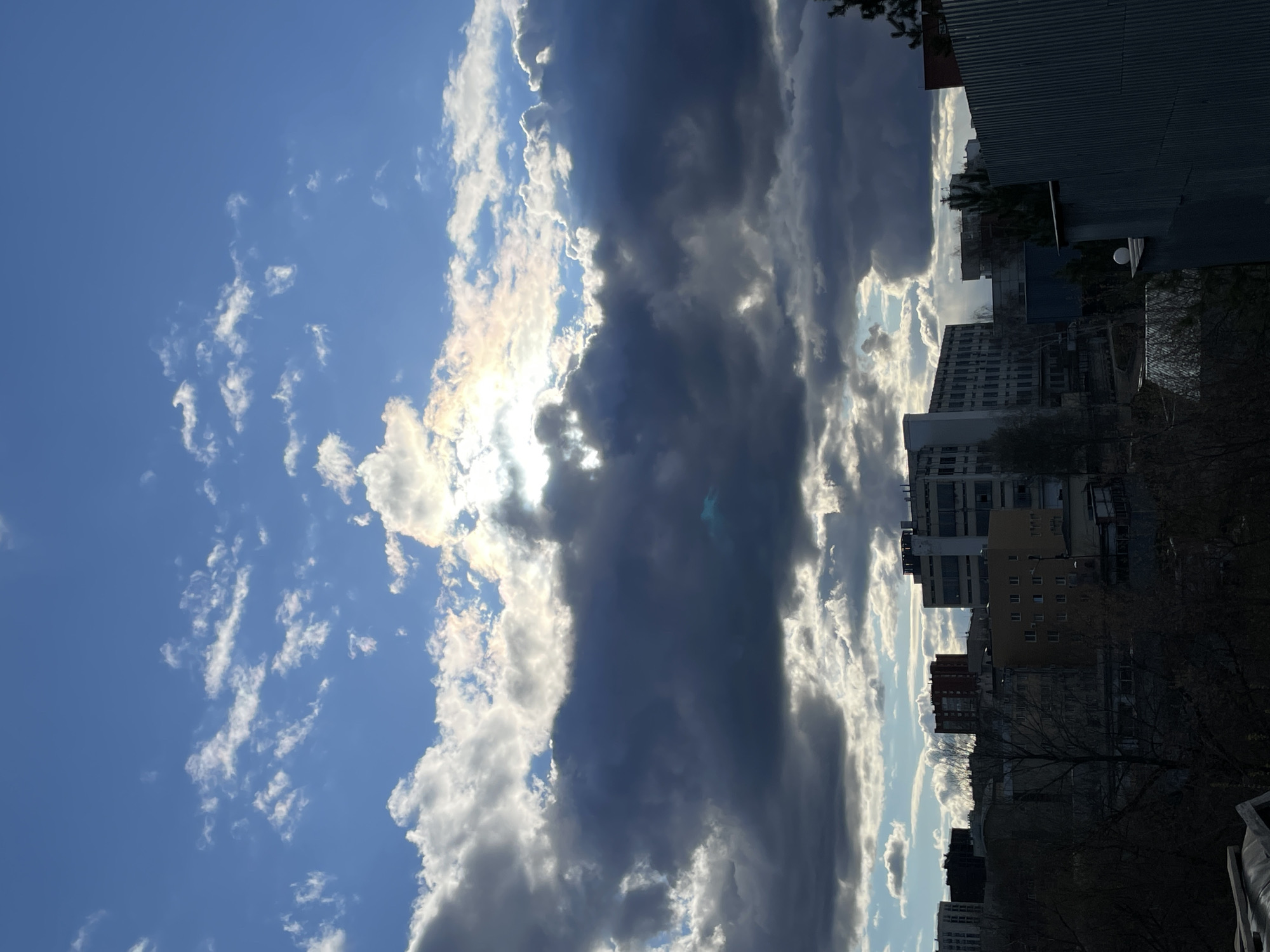 The image depicts an urban sky dominated by dynamic cloud formations and the upper parts of city buildings. A prominent, dark band of clouds stretches horizontally across the middle of the frame, contrasting sharply with patches of bright blue sky and sparser white clouds visible in the upper left. Intense sunlight pierces through a gap within the dark clouds on the left side, creating a brilliant, almost iridescent glare that vividly illuminates the edges of the surrounding white clouds.
On the right side of the image, the upper sections of various multi-story urban buildings are visible. These structures, likely a mix of residential and commercial, are largely silhouetted or cast in shadow due to the backlighting from the sun. In the lower right foreground, the bare, leafless branches of deciduous trees can be seen, suggesting a cooler season.
The strong, low-angle light peeking through the clouds indicates either early morning or late afternoon. The dramatic interplay between the heavy, dark clouds and the intense sunlight creates a striking visual effect, hinting at a day with rapidly changing weather. No discernible human activity is taking place, and there is no visible text. The scene offers a view of a city sky, characteristic of an urban environment such as Izhevsk, Russia.