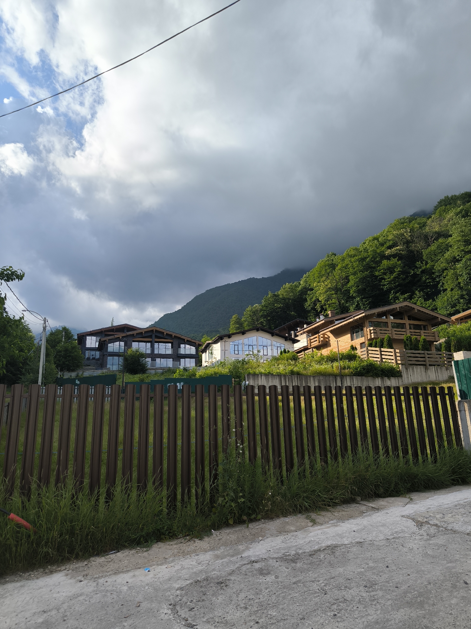 The image captures a scenic view of modern wooden houses nestled on a hillside in Krasnaya Polyana, Russia. The houses are built in a contemporary style, featuring large windows and natural wood exteriors. They are situated against a backdrop of lush green forested mountains, with dramatic, heavy clouds dominating the sky, suggesting an approaching storm or recent rainfall. In the foreground, a tall, vertical wooden fence runs across the image, separating the viewer from the properties. The ground in front of the fence is paved with a rough, grey concrete surface, and wild grass grows along the base of the fence. The overall impression is one of a remote, upscale residential area surrounded by natural beauty, with the weather adding a touch of dramatic atmosphere.