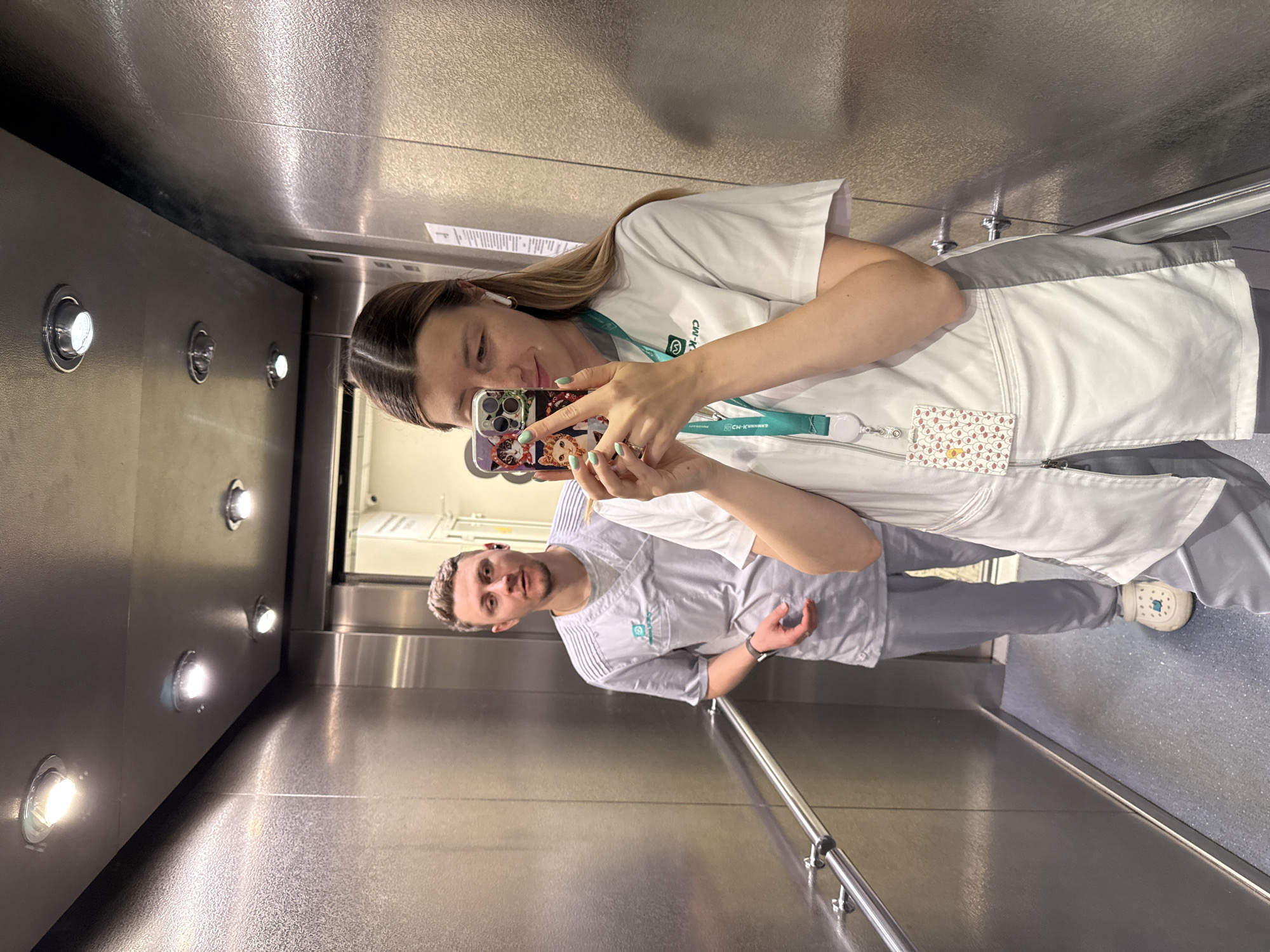 Two individuals, a woman and a man, are captured in a mirror selfie taken inside a metallic elevator in Moscow, Russia. The image is oriented sideways, with the subjects appearing to stand on the left wall.
The woman, positioned in the foreground and appearing to take the selfie, is holding a smartphone with a distinctive cat-themed case. She has long, light brown hair, is wearing an earbud in her left ear, and has mint green nail polish. Her attire consists of a white and light grey medical or professional uniform (scrubs) with a visible logo that appears to be "CW-K..." and a lanyard featuring the text "СШ-КУННИКИ". A colorful patterned ID badge is attached to her lanyard.
Behind her, the man stands with a neutral expression, looking towards the camera. He has short, light-colored hair and is wearing a light grey medical or professional uniform with textured patterns on the shoulders and a similar "CW-K..." logo. He wears a dark watch on his left wrist and light-colored clogs with a blue design on his feet, typical of medical footwear.
The elevator interior features polished metallic walls and floor, creating reflections. Several small, circular light fixtures are visible along the walls, casting bright illumination. A chrome handrail runs along the lower part of the right wall (as viewed in the sideways orientation). The overall scene suggests a professional environment, likely a hospital or clinic, given the attire of the subjects.