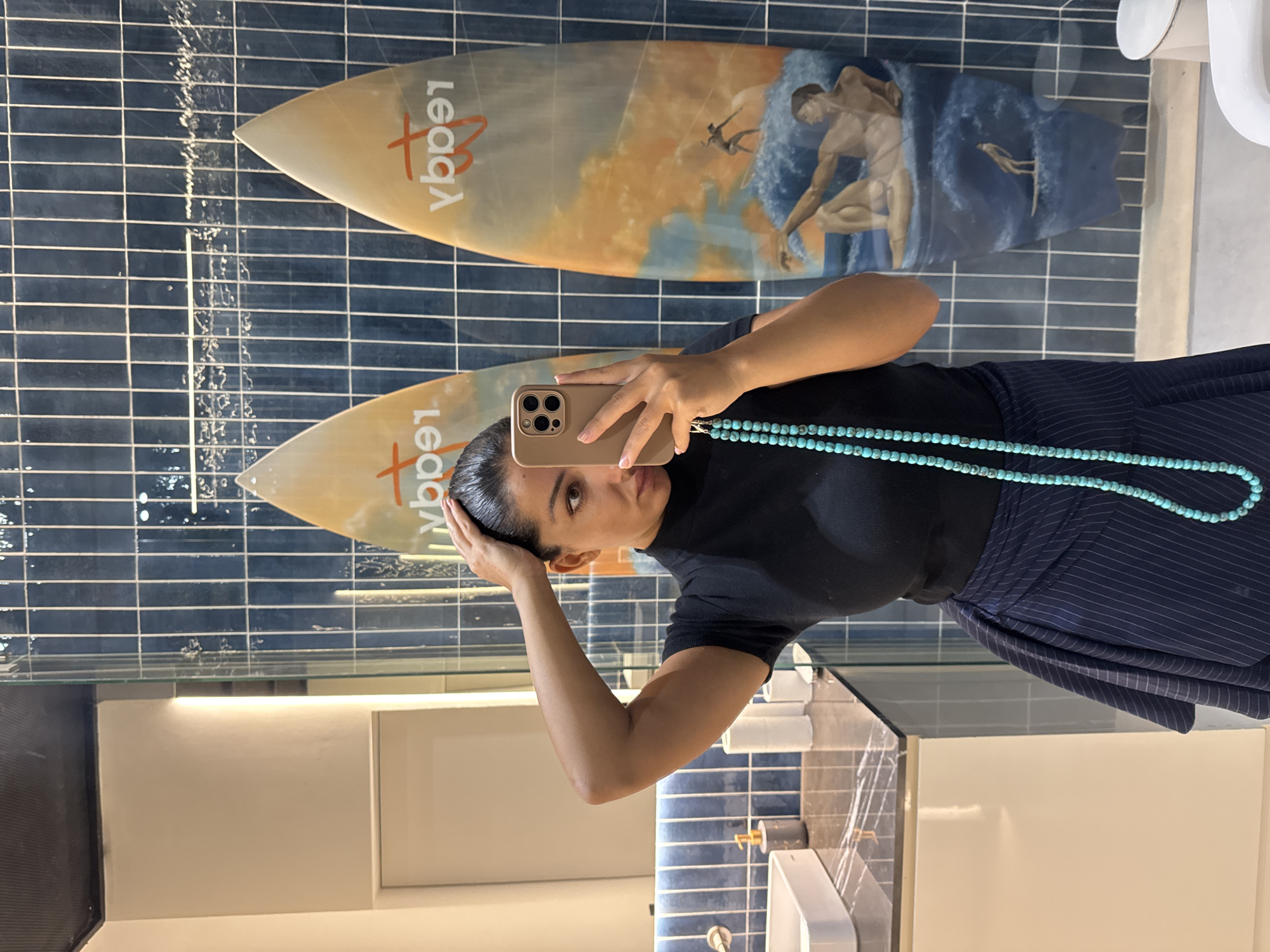 A person is taking a selfie in a restroom. The restroom walls are lined with dark blue tiles. Two surfboards are mounted on the wall as decoration. The surfboards have the word "Legacy" written on them, and one depicts a painting of a man in the water. The person is wearing a dark-colored top and a blue and white striped skirt. A turquoise beaded necklace is around their neck. A sink and counter are visible in the lower part of the frame. Based on the surfboards and overall aesthetic, it's likely the photo was taken at a location in Kuta Selatan, Indonesia.