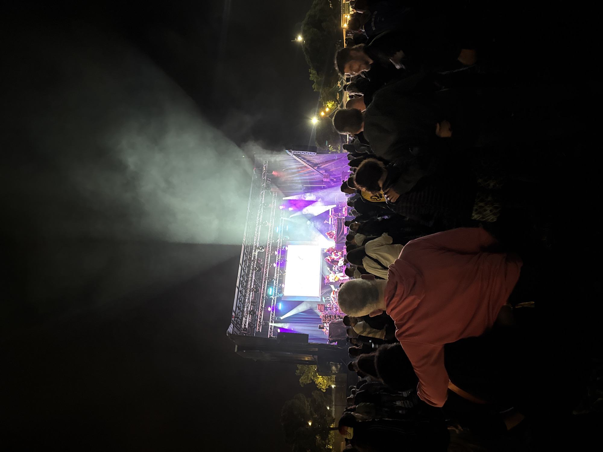 The image shows a nighttime outdoor concert with a stage illuminated by bright lights and smoke. A crowd of people is gathered in front of the stage. The concert appears to be in an urban setting, as there are trees and streetlights visible in the background.
Based on the location, and the nature of the image, it is likely this scene is from an outdoor concert during the Festa Major de Vilanova i la Geltrú in Spain, possibly at Parc de la Torre d'En Dolcet where many outdoor concerts are held.