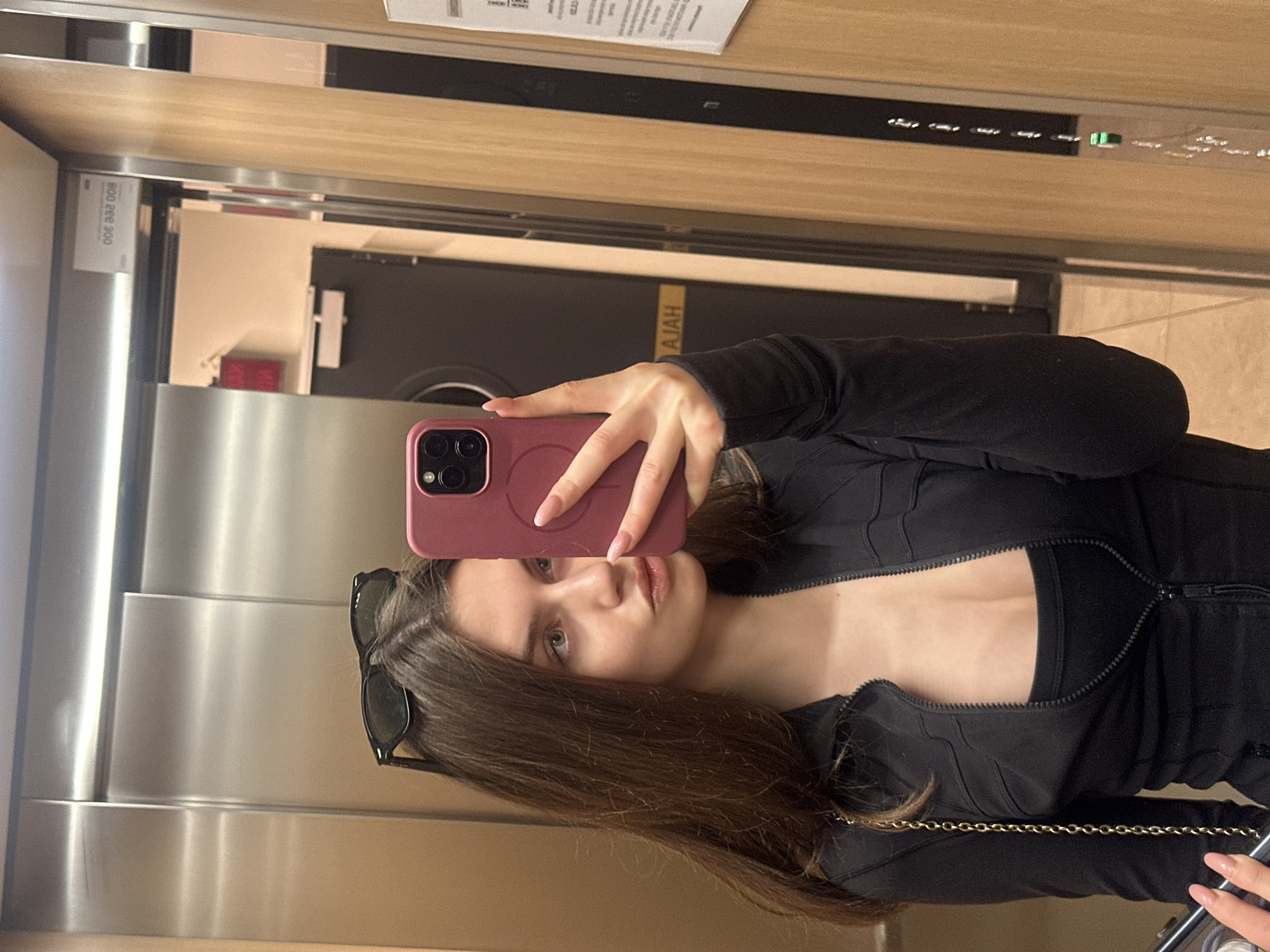 A woman is taking a selfie in an elevator with a pink phone. She is wearing a black top with a zipper and has sunglasses perched on her head. The elevator has stainless steel walls and a wooden panel above the mirror. A door is visible in the reflection, with text “AJAH”. Above the door there is a printed information sheet.