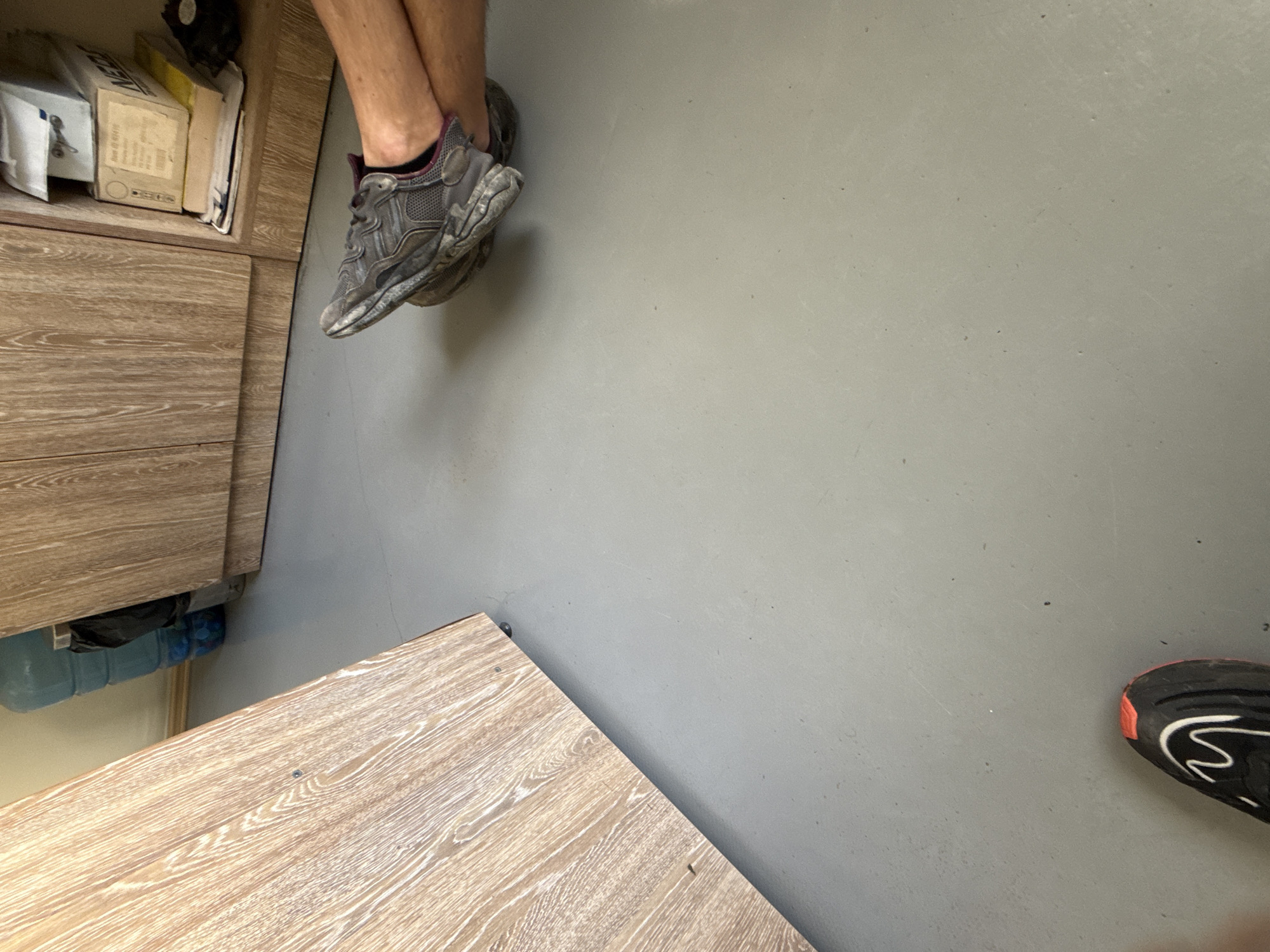 The image shows a section of a room with light gray flooring. There is a wooden cabinet or storage unit to the left, holding items including books and packaged goods. A person is seated with their legs crossed, wearing gray and maroon athletic shoes. In the bottom right corner of the image, there is another athletic shoe, black with a red accent and white design. The location is most likely indoors, but there is no indication of it being in Stara Zagora, Bulgaria.