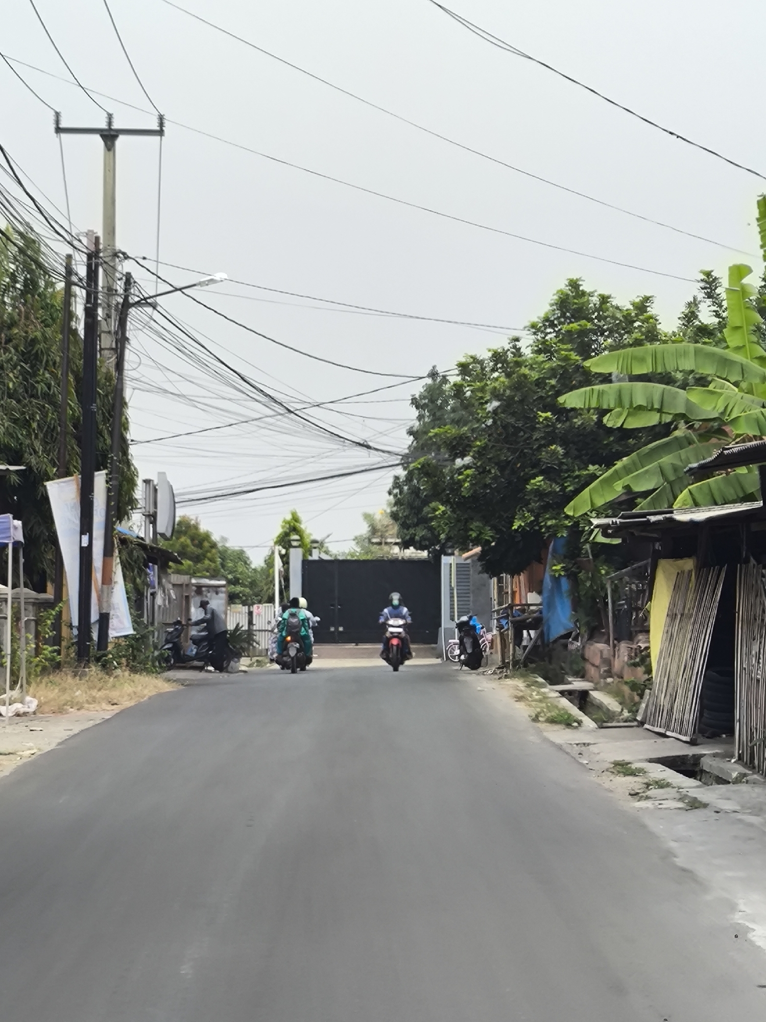The image shows a street scene in Cilegon, Indonesia, under an overcast sky. The foreground is dominated by an asphalt road. In the middle ground, two individuals are riding motorcycles. The person on the left is riding a teal motorcycle, and the person on the right is riding a red motorcycle, both with helmets on. Several other motorcycles are parked along the side of the road. The left side of the street features a tall utility pole with numerous wires, trees, and a gray fence with banners. The right side of the street has a narrow sidewalk and drainage channel, with a building constructed of wood and corrugated metal, adorned with hanging tarps and stored wooden planks. In the background, a large black gate is visible, suggesting an entrance to a property. The overall environment appears to be a residential or mixed-use area with a utilitarian aesthetic.