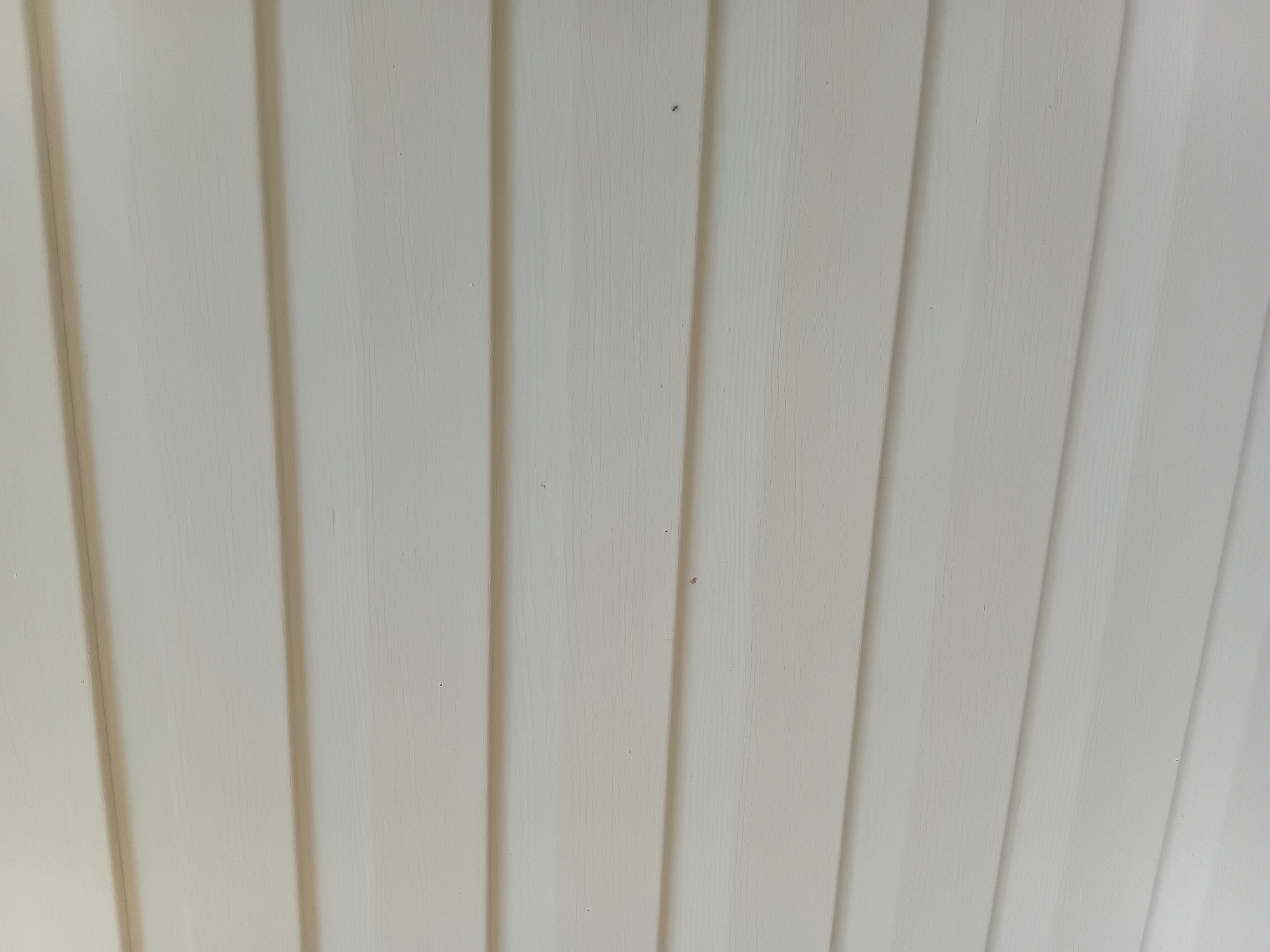 The image shows a close-up view of a light-colored, textured surface, likely siding or paneling. Vertical lines create a ribbed or grooved effect, with subtle shadows indicating a raised or recessed pattern. The surface appears to be uniformly colored, possibly off-white or a very pale beige. There are a few small, dark specks scattered across the surface, which could be dust or minor imperfections. The lighting suggests it is daytime, but there are no other environmental cues like windows or outdoor scenery. The image is an abstract depiction of a building material, focusing on its texture and form. There is no discernible activity or event taking place, and no people or identifiable objects are present. The location context of Verkhniachka, Ukraine, does not provide specific visual information within the frame itself.