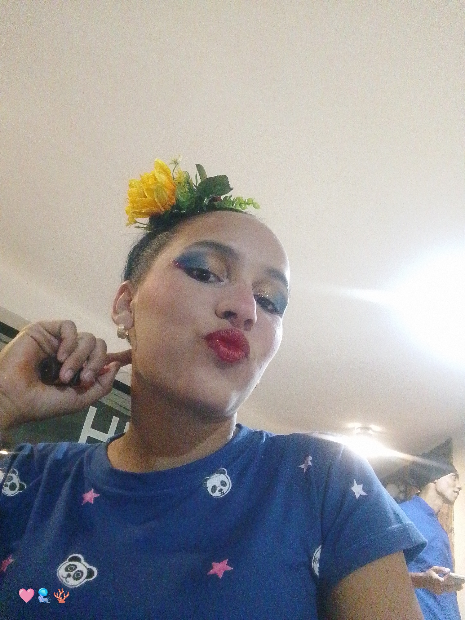 A woman is posing for a selfie in an indoor location, possibly a home or a shop, in the city of Unknown, Unknown. She is wearing a blue t-shirt with panda and star patterns. She has a yellow flower and green leaves hairpiece. Her makeup includes blue eyeshadow with glitter and red lipstick. She is holding a small stick to her ear and making a kissing face. A blurry male figure is partially visible on the right side, wearing a blue shirt and a black beanie. The background includes a white ceiling.