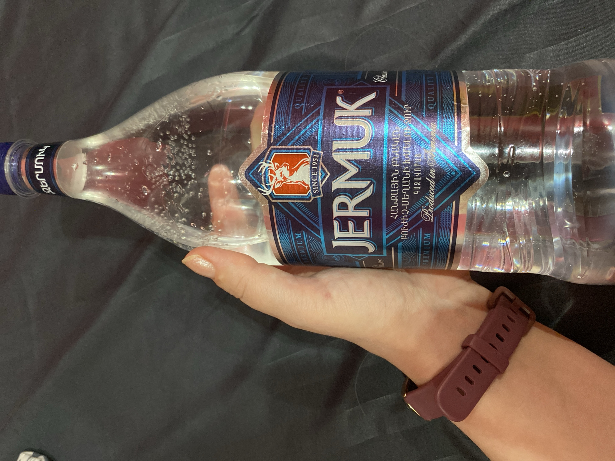 A hand wearing a burgundy watch band is holding a clear plastic bottle of Jermuk mineral water. The bottle has a blue and silver label with text in Armenian and English, including "JERMUK", "SINCE 1951", and "Produced in Armenia". The bottle is filled with water containing numerous small bubbles. The background is a dark, textured surface. There is no indication of the city of Yerevan in the image.