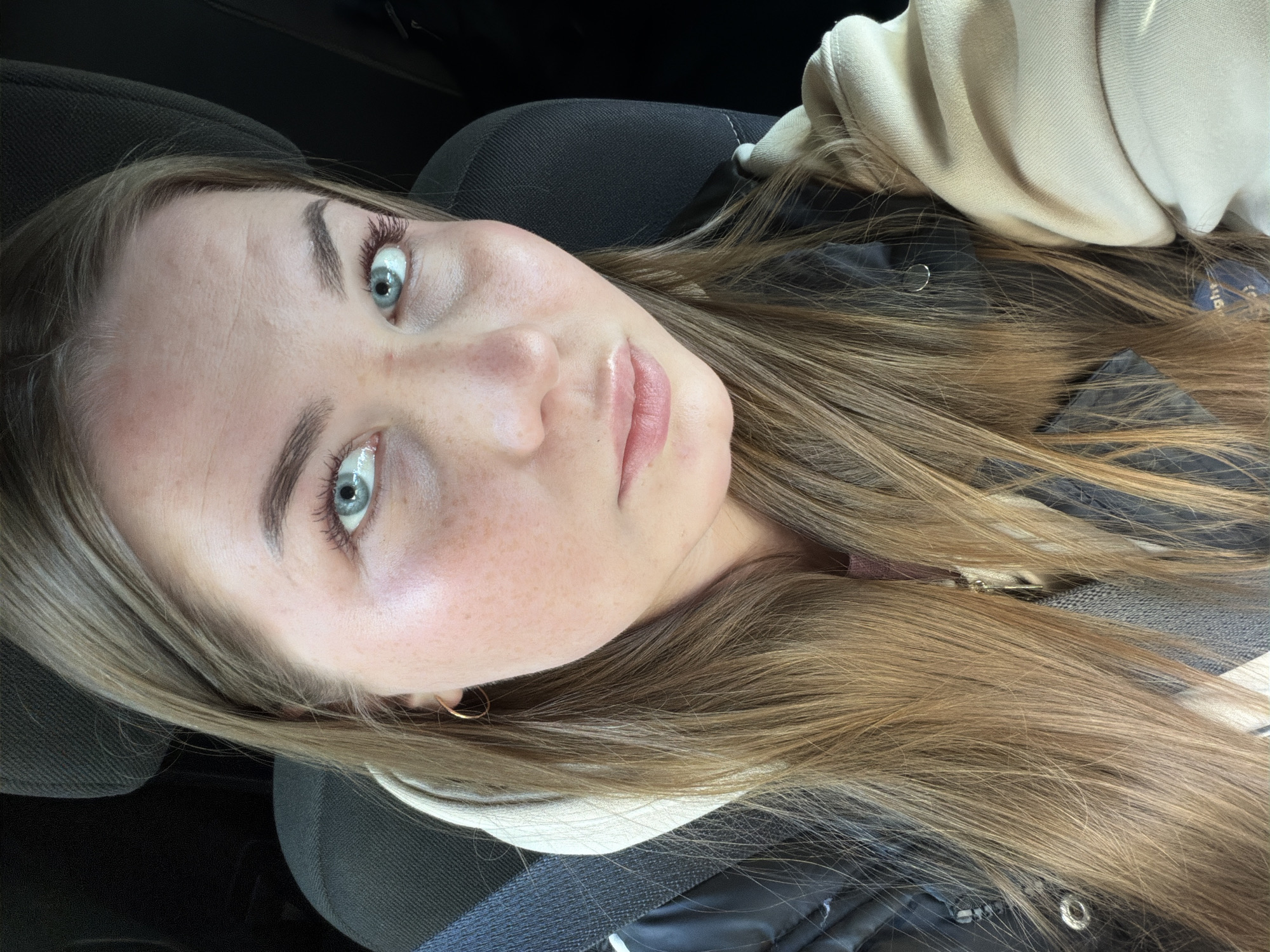 A young woman with fair skin and light brown hair is positioned in what appears to be the front passenger seat of a car. She is wearing a light-colored hoodie, a dark jacket, and has a seatbelt across her torso. Her eyes are a striking blue-green color, and she has freckles on her face. She is looking directly at the camera. The background suggests the interior of a vehicle. This image does not provide information about the city of Chaikovsky, Russia.