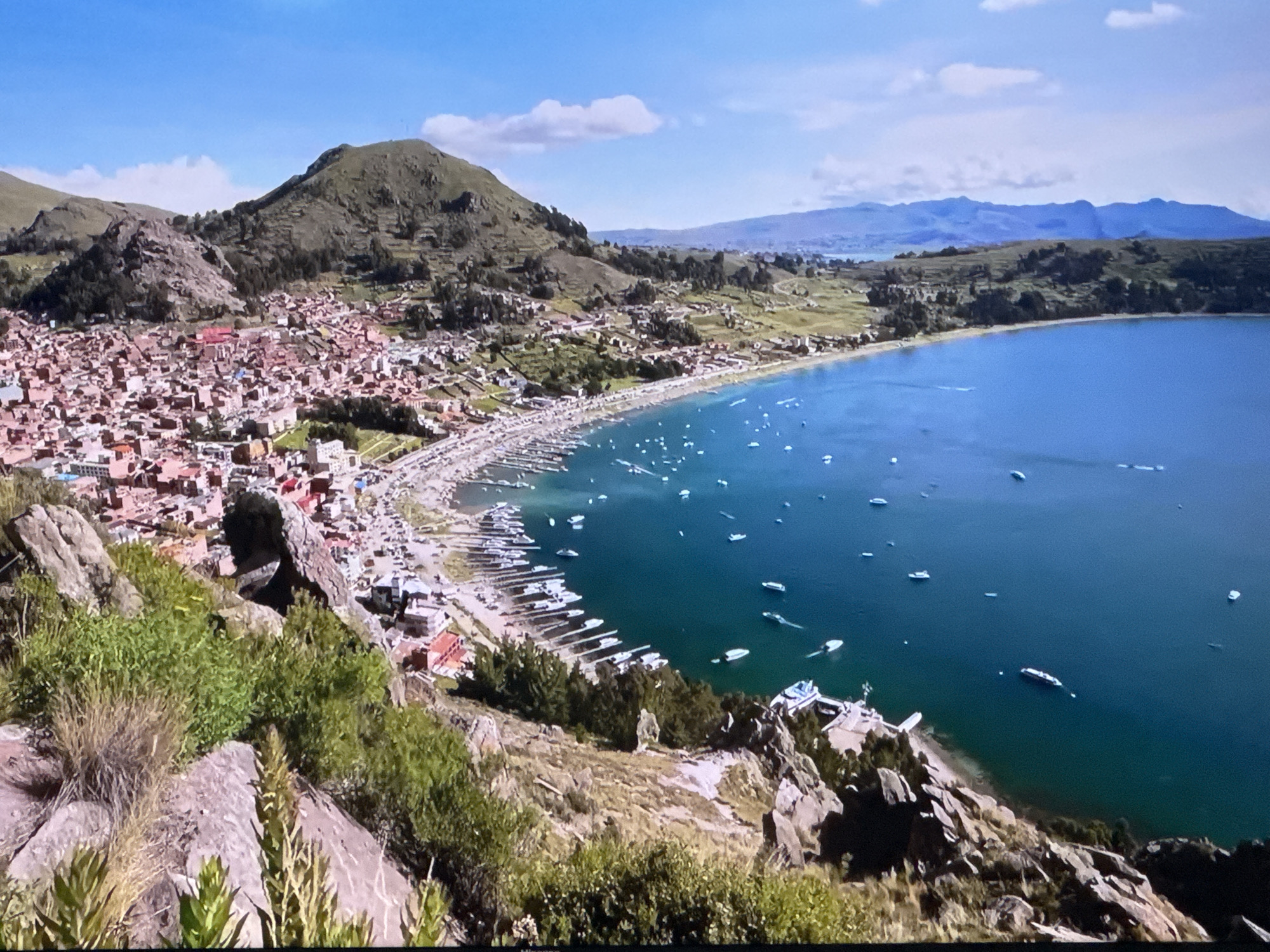 The image shows a panoramic view of a coastal city nestled at the base of rolling green hills, with a vast body of turquoise water stretching to the horizon. The city, characterized by its densely packed buildings with red-tiled roofs, extends along the shoreline. Numerous boats are visible in the water, both docked along the coast and scattered across the open expanse. The foreground is dominated by rocky terrain with sparse vegetation, suggesting a natural, somewhat rugged landscape overlooking the developed area. In the distance, a range of hazy blue mountains forms the backdrop against a bright sky with scattered clouds. The scene appears to be captured from an elevated viewpoint, providing a sweeping perspective of the city, its harbor, and the surrounding natural environment.