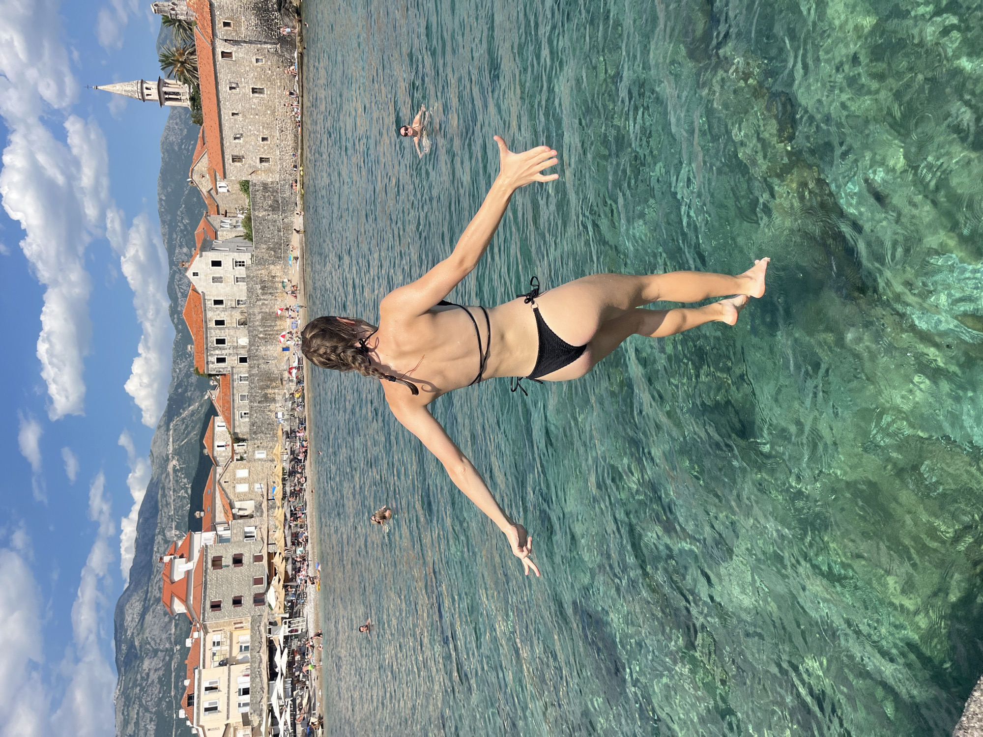 A woman in a black bikini leaps into clear turquoise water, her arms outstretched. In the background, a picturesque town with red-tiled roofs and stone buildings rests against a backdrop of mountains. People sunbathe on the beach, and others swim in the sea. The water is so transparent that the rocky seabed is visible beneath the surface. The sky above is a brilliant blue, dotted with fluffy white clouds.