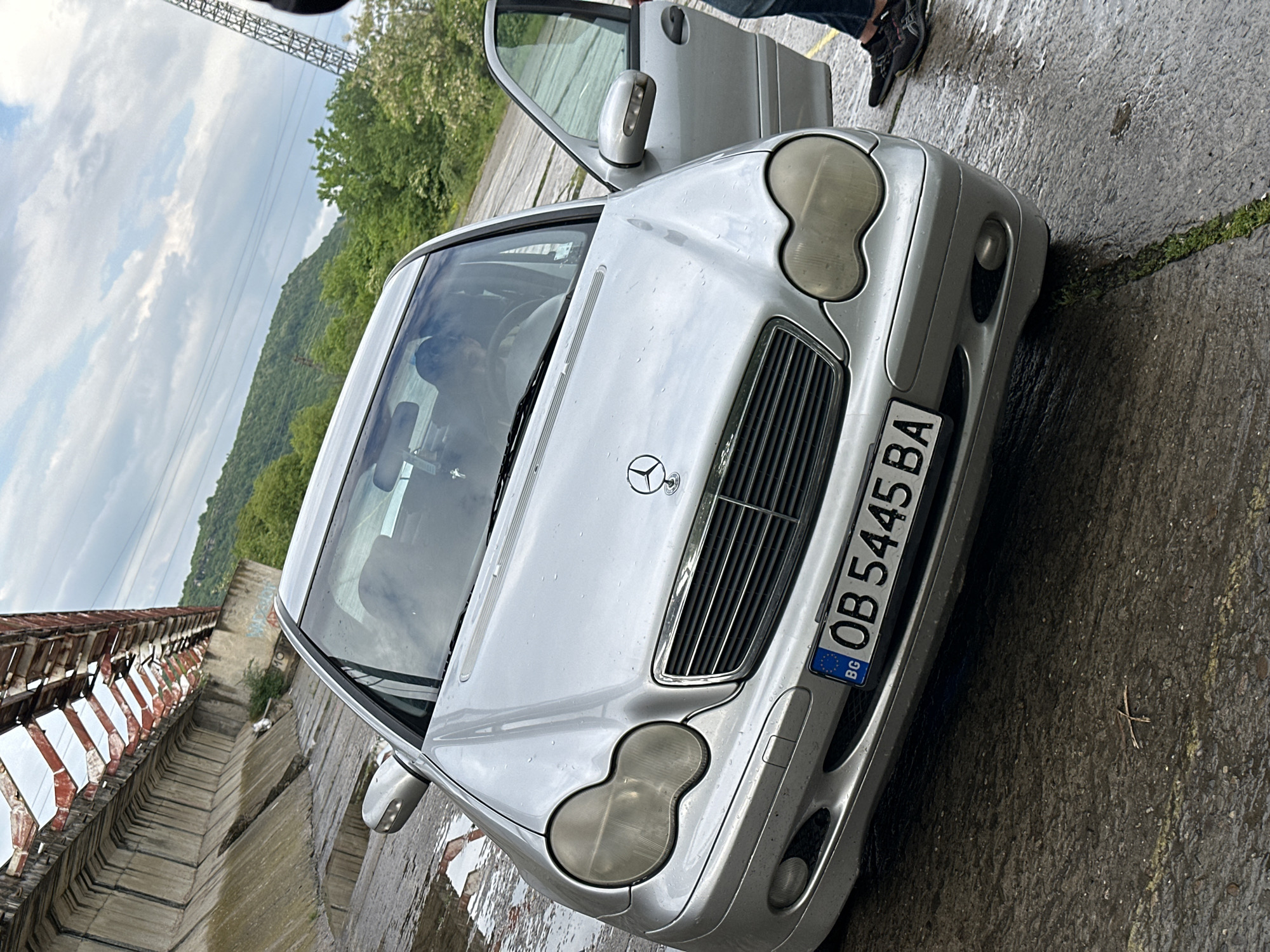 The image shows a silver Mercedes-Benz sedan, displaying Bulgarian license plate OB 5445 BA, parked on a concrete surface. The car's driver-side door is open, revealing a glimpse of the interior and a person standing nearby. The car appears to be wet, suggesting recent rain. In the background, there's a structure with red and white elements and vegetation. In the distance, there is a hillside with trees. Based on the presence of Bulgarian license plate and the location specified in the instructions, the photo was taken in Umarevtsi, Bulgaria.