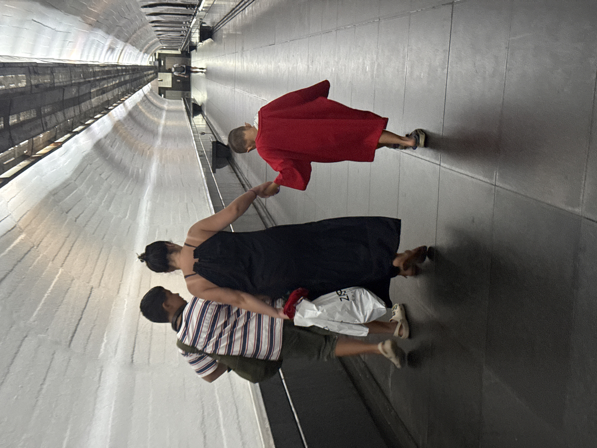 In an enclosed, arched passageway, possibly a pedestrian tunnel or underpass in Yalta, Ukraine, three individuals are walking away from the camera. On the left, a male, appearing to be a child or young teenager, wears a striped t-shirt and shorts, with a dark bag slung over his shoulder. Next to him, a woman, likely his mother, is dressed in a long, black, spaghetti-strap dress. She is holding a white shopping bag with red items inside. On the right, a smaller child, also male, is wearing an oversized bright red cape-like garment and is holding the woman's hand. The passageway has white, textured walls and a dark floor composed of large square tiles. The ceiling is visible in the background, appearing to be a metal structure with overhead lighting. The overall lighting is dim, with light sources creating shadows on the floor.