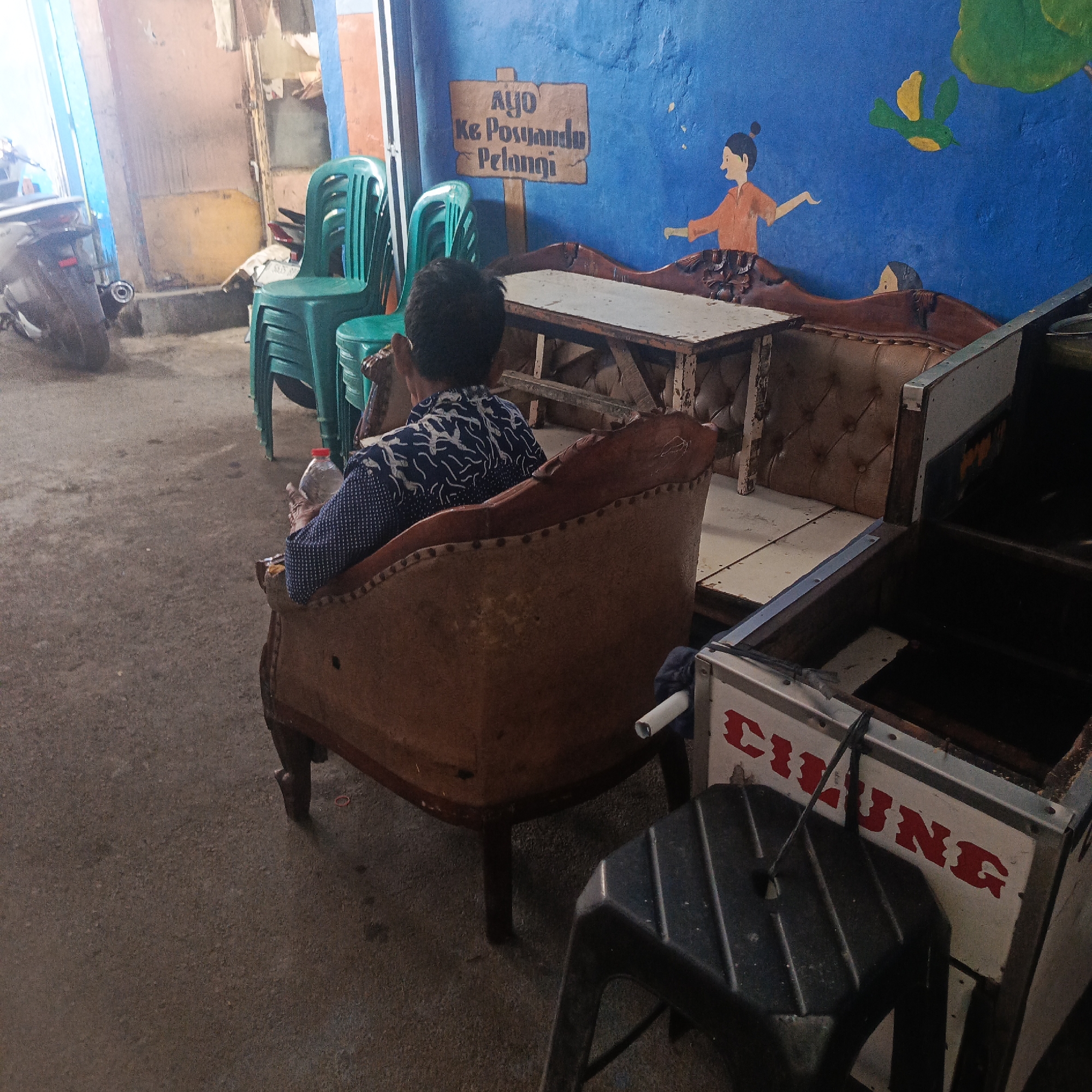 In a rustic outdoor setting, a person sits in a cushioned chair, facing away from the camera, holding a plastic bottle. The environment appears to be a casual waiting area or a small shop, possibly a food stall, with an open-air feel. In the background, green plastic chairs are stacked, and a motorcycle is parked to the left. A mural adorns the blue wall, featuring a child-like figure and the text "AYO KE POSYANDU PELANGI," which translates to "Come to Pelangi Posyandu" (Posyandu is a community health post in Indonesia). A wooden table sits in front of a tufted brown sofa. To the right, a food cart with the word "CIUNG" visible in red lettering is partially in frame, suggesting a commercial establishment. The ground is a rough concrete surface. This scene is located in South Jakarta, Indonesia.