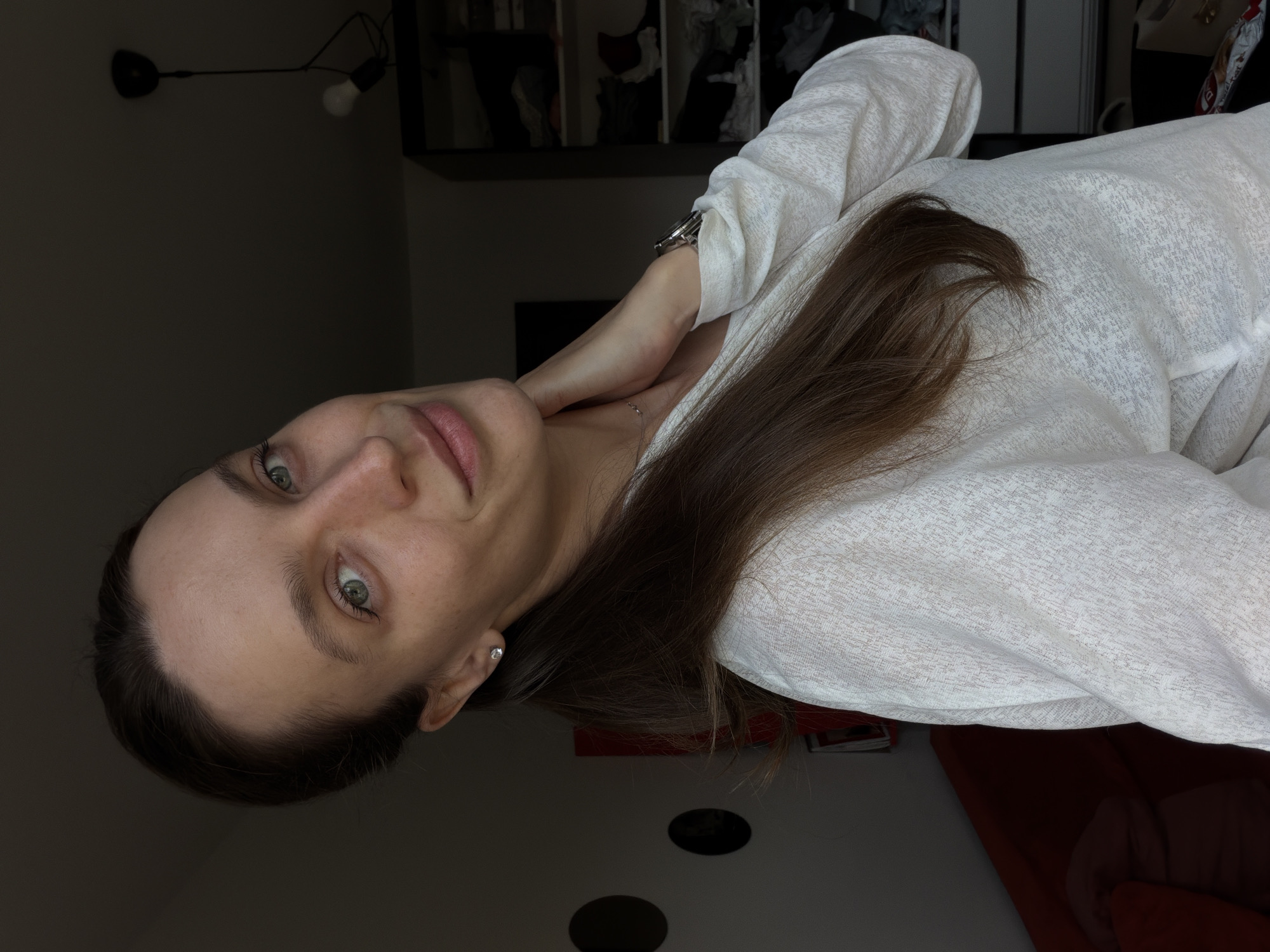 A woman with long, brown hair and green eyes is pictured indoors. She is wearing a light-colored shirt. Her left hand is near her neck. A wristwatch is visible on her left wrist. A light fixture and shelves with various objects are in the background. The scene appears to be dimly lit. The location in the city of Vozdovac Urban Municipality, Serbia cannot be determined from the image.