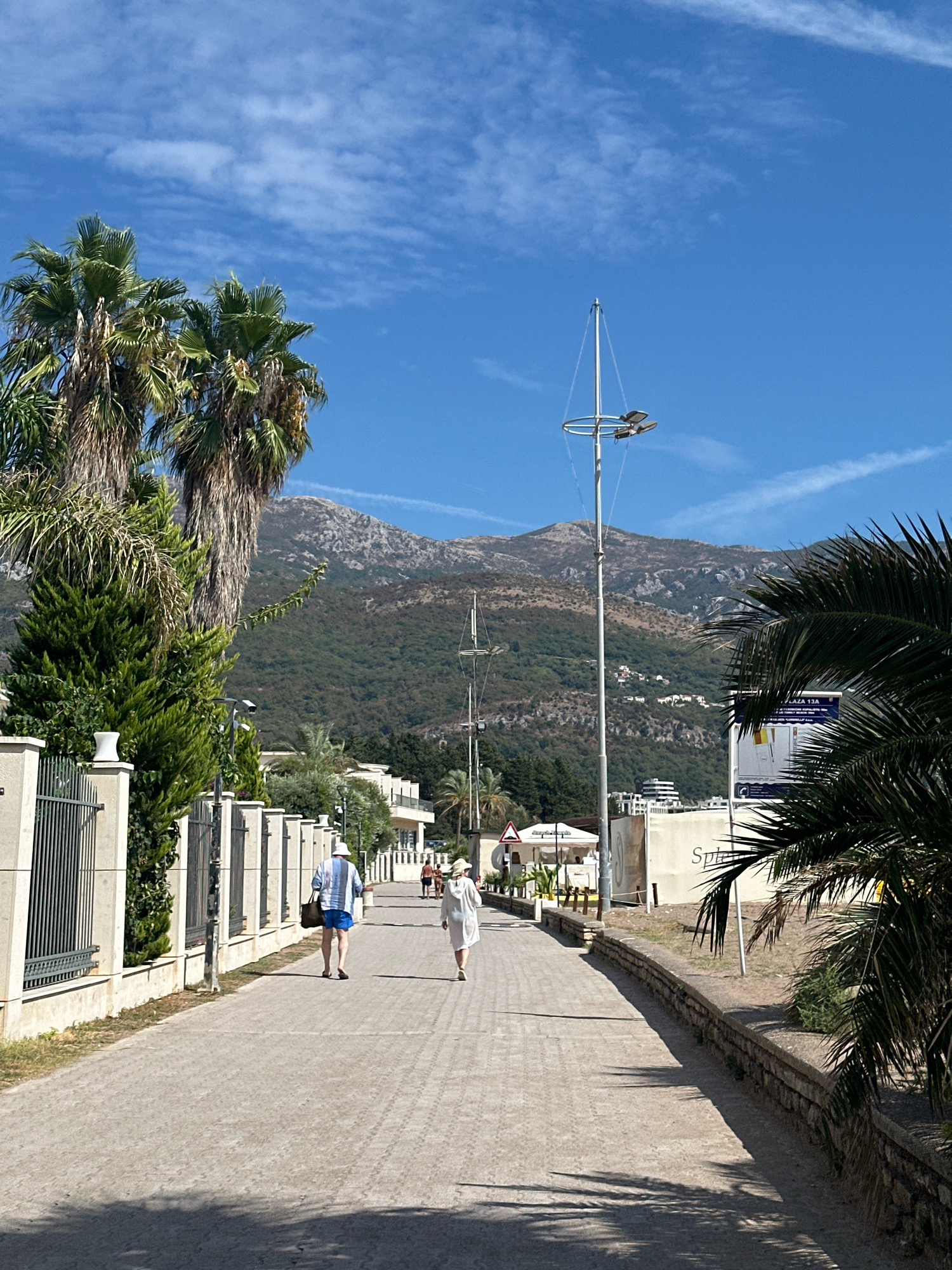 The image shows a street in Boreti, Montenegro, with two people walking away from the camera on a paved path. The path is lined with a low stone wall on the right and a metal fence with greenery behind it on the left. In the background, there are buildings and lush green mountains under a bright blue sky with scattered clouds. Palm trees are visible on the left and right sides of the image. A tall pole with lights is also present. The overall scene depicts a sunny day in a coastal area.