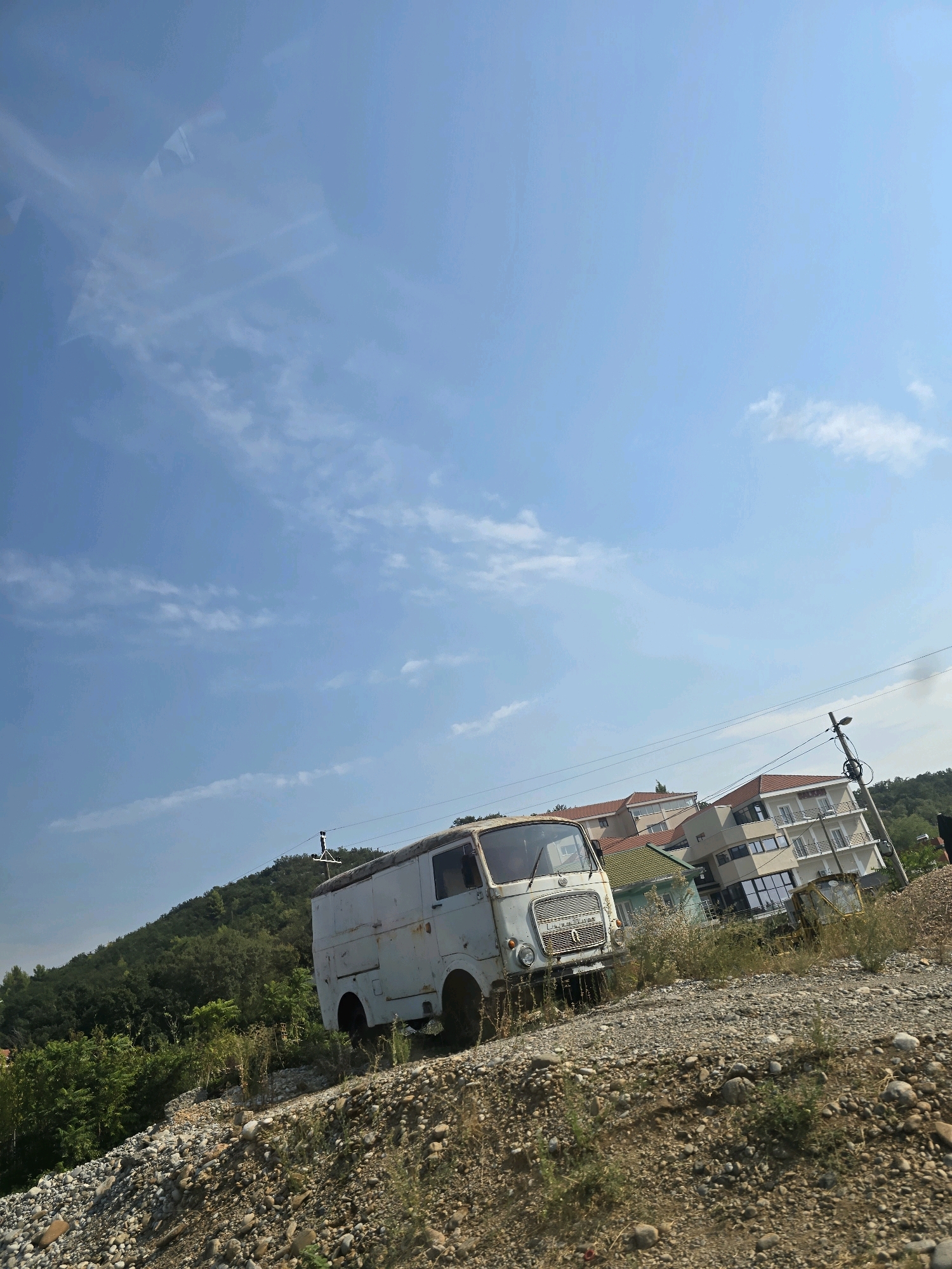 An abandoned white van sits on a rocky, overgrown incline in a semi-urban setting. Behind the van, several multi-story buildings with red-tiled roofs are visible, suggesting a residential or commercial area. A utility pole with wires stands to the right of the buildings. A lush green hillside with dense trees rises in the background, contrasting with the developed structures and the neglected vehicle. The sky is a clear blue with a few wispy clouds. The scene appears to be located in or near Podgorica, Montenegro, with the van likely positioned on the outskirts of the city, possibly near a construction site or an undeveloped area. There are no people clearly visible in the image.