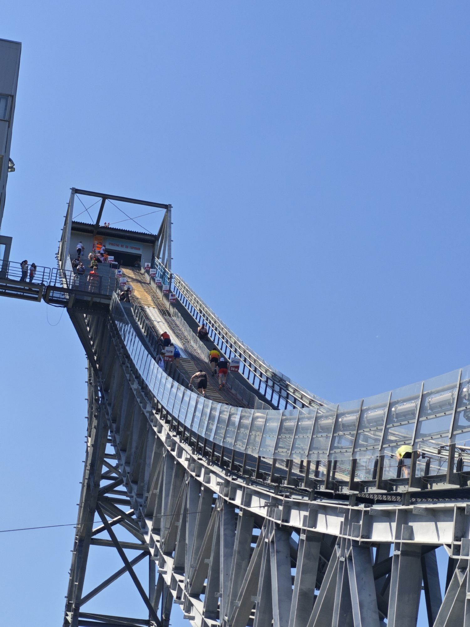 The image shows the ski jumping complex "Flying Ski" in the city of Прикамский, Russia. The complex is constructed of gray steel beams and enclosed with transparent panels. People are present in the image, both on the upper platform near a structure with an overhang and walking up the sloped ramp. The sky is clear and blue.