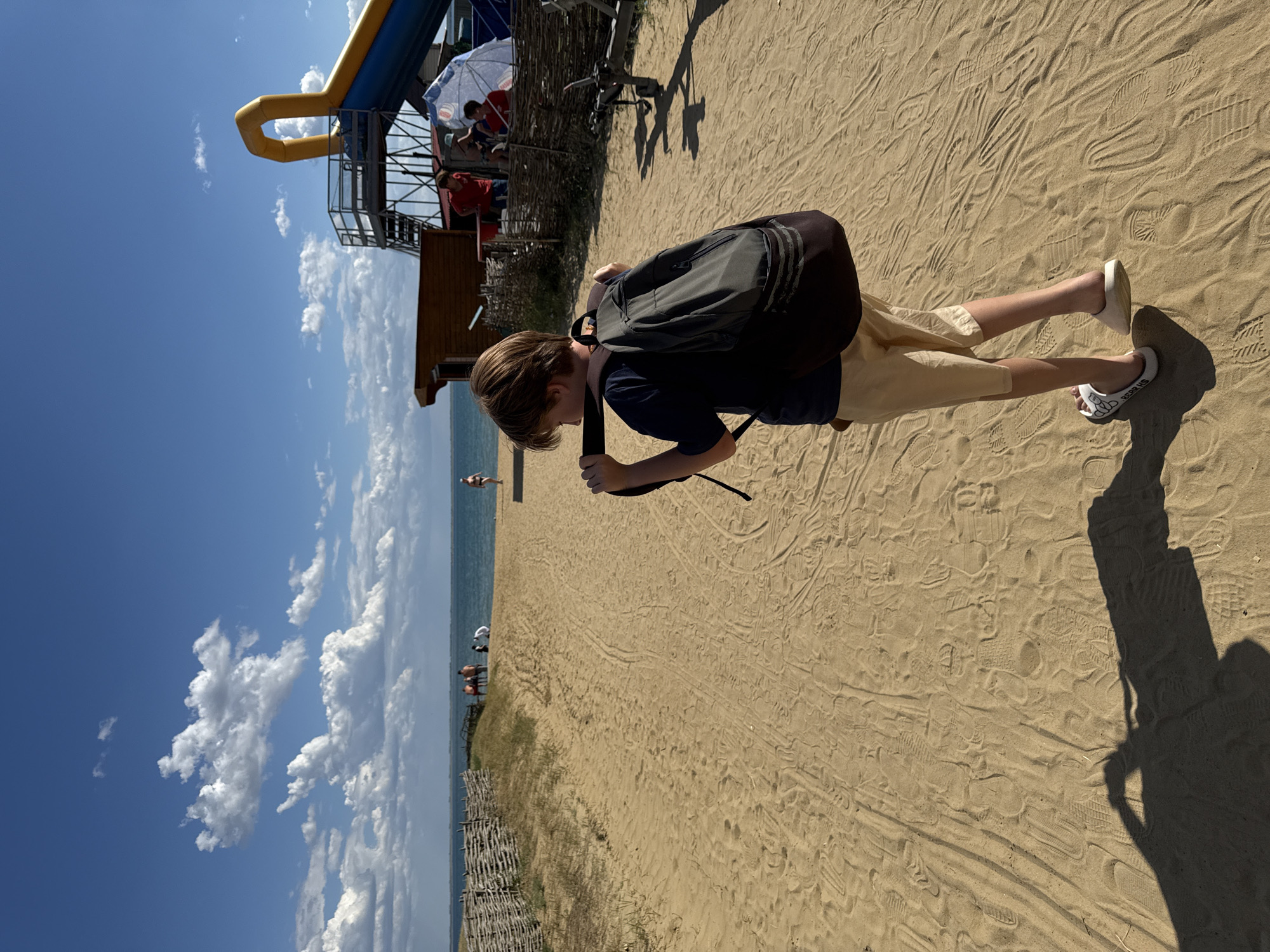 The image depicts a sandy path leading towards a body of water, likely a sea or lake, under a blue sky with scattered white clouds. On the right side of the path, there is an inflatable water slide structure with a yellow tube and a blue slide, along with a small wooden building. Several people are visible in the background near the water and the slide area. A young person with a backpack is walking away from the camera on the sandy path. The environment appears to be a recreational area, possibly a beach or amusement park, in Yarovoye, Russia, given the presence of the water slide and the general landscape. The ground is covered in sand with numerous footprints.