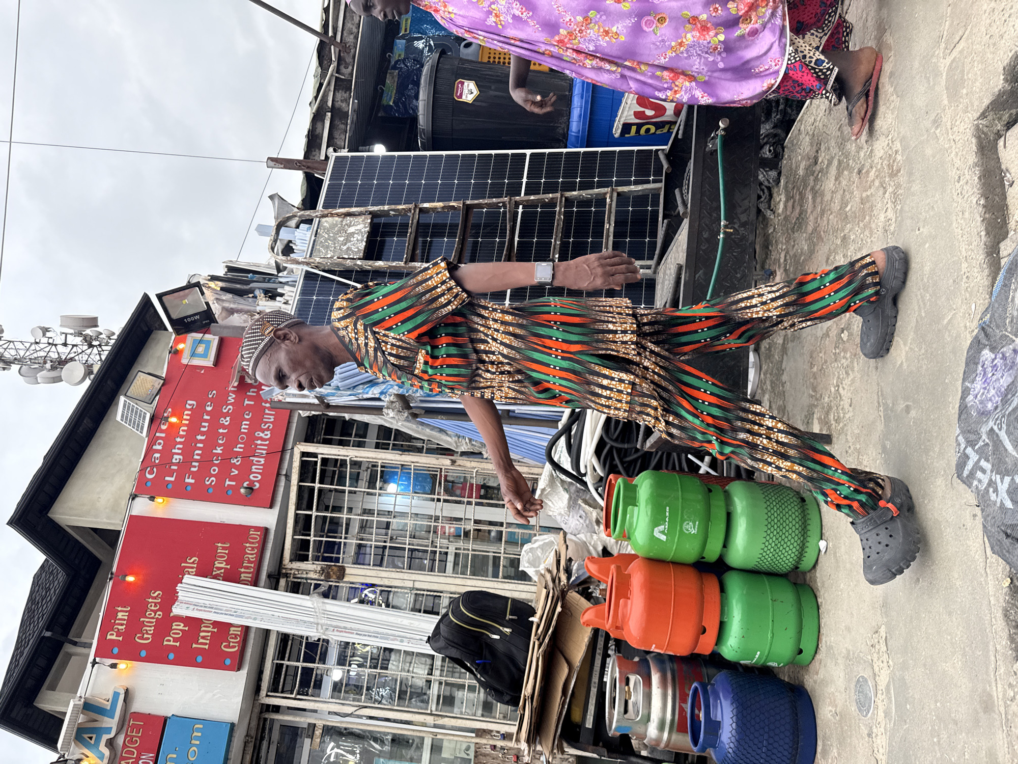 In Ado, Nigeria, a man in a patterned dashiki and pants walks past a store. The store has signs listing various products and services including "Paint," "Gadgets," "Import Export," "Cables," "Lightning," "Furnitures," "Socket & Swi," and "Tv & Home Th Conduit & surf." Adjacent to the store are stacks of propane tanks, one blue, two orange, and two green. A ladder leans against a solar panel array. The man is wearing black crocs. To the right, a woman in a purple floral dress and a pink headscarf walks away from the camera. She is wearing pink flip-flops. The ground is concrete.